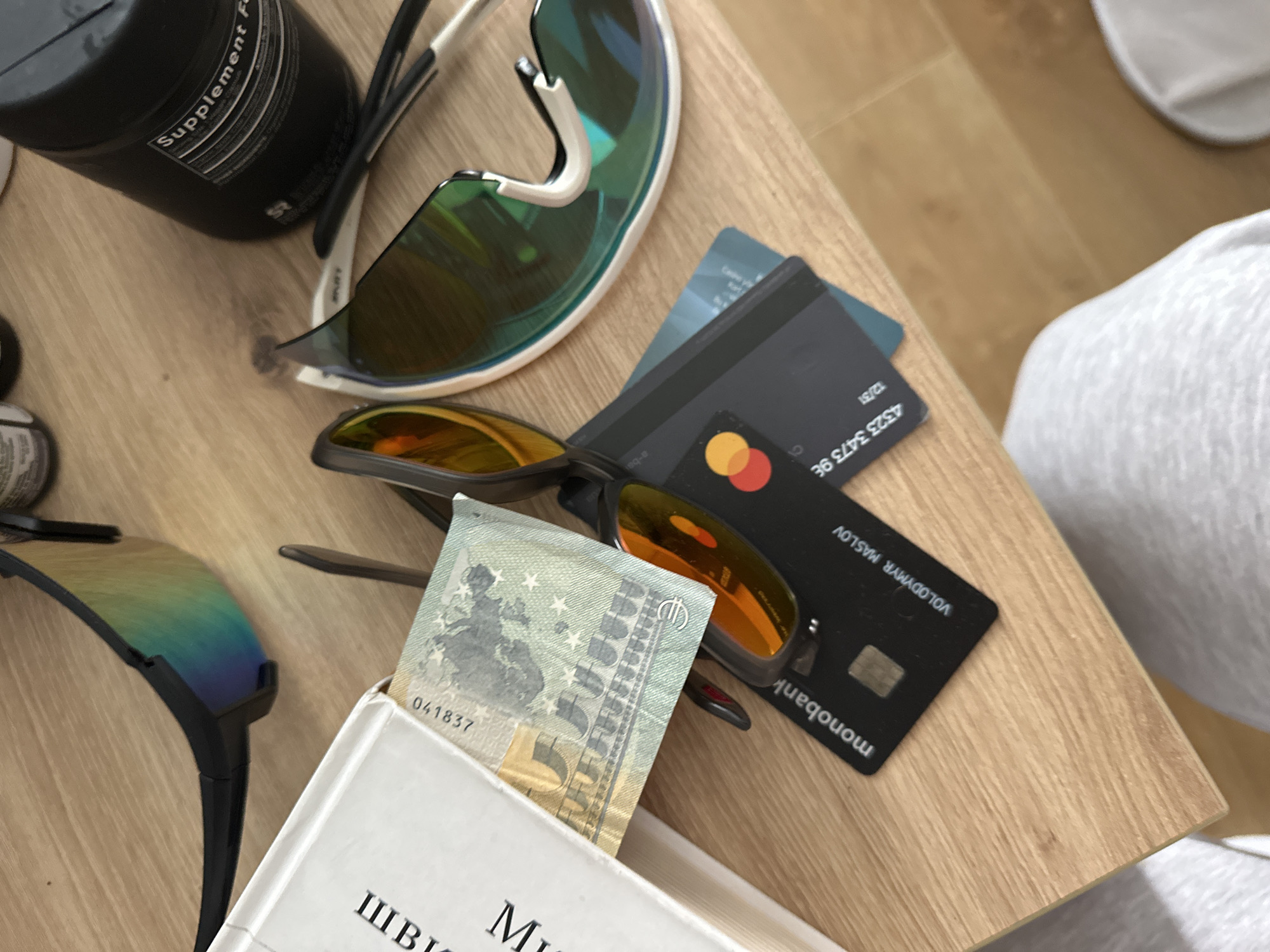 The image shows a close-up, overhead view of a wooden table surface, likely in an indoor setting. The lighting suggests a bright environment, possibly from natural light.
On the table, several objects are arranged. In the foreground left is a portion of a white book, with the beginning of some text visible. Tucked into the book is a Euro banknote, appearing to be a 5 Euro note, with the serial number "041837" visible.
To the right of the book and banknote are two pairs of sunglasses. One pair, with black frames and iridescent lenses, lies horizontally across the top of the frame. Another pair, with white frames and mirrored, multi-colored lenses, is positioned to the right and slightly below the first pair.
Beneath the sunglasses, and slightly overlapping, are two black credit cards. The card in the foreground has the logo "monobank" and the name "VOLODYMYR MASLOV" printed on it. The other card is partially obscured.
In the upper right corner of the image, a black container, possibly a supplement bottle, is visible. The label on the bottle partially reads "Supplement F".
The context of Donji Pobori, Montenegro, is not directly evident from the visual elements within the image itself. The scene depicts everyday personal items on a table, lacking any specific indicators of a particular city or location.