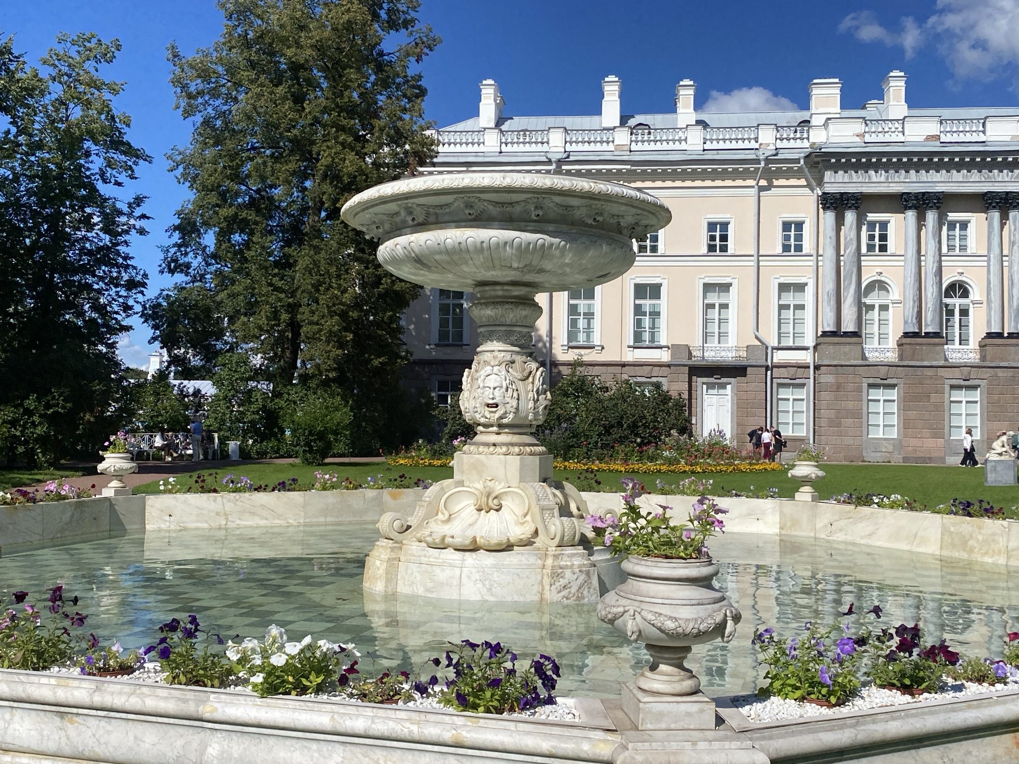 This is a photograph taken outdoors on a sunny day in Pushkin, Russia, likely within the grounds of the Catherine Palace. In the foreground, there is a large, multi-tiered marble fountain with intricate carvings, including faces and decorative motifs. The fountain is filled with clear water, and its basin is lined with a checkered pattern. Around the perimeter of the fountain, there are marble borders filled with white pebbles and various potted flowers in shades of purple, pink, and white. Two ornate urns, one on the left and one on the right edge of the fountain, also contain potted plants.
In the mid-ground, behind the fountain, is a lush green lawn. Beyond the lawn, a light-pink classical-style building with white trim and multiple windows is visible. The building features a prominent portico with large columns. Trees with dense green foliage are present on the left side of the image, and more greenery and flower beds are scattered throughout the scene.
In the background, several people can be seen walking on the lawn and near the building. A few figures are gathered in groups, some appearing to be tourists. The sky is a bright, clear blue with a few scattered white clouds. The overall scene depicts a formal garden setting with a grand fountain and a large palace.