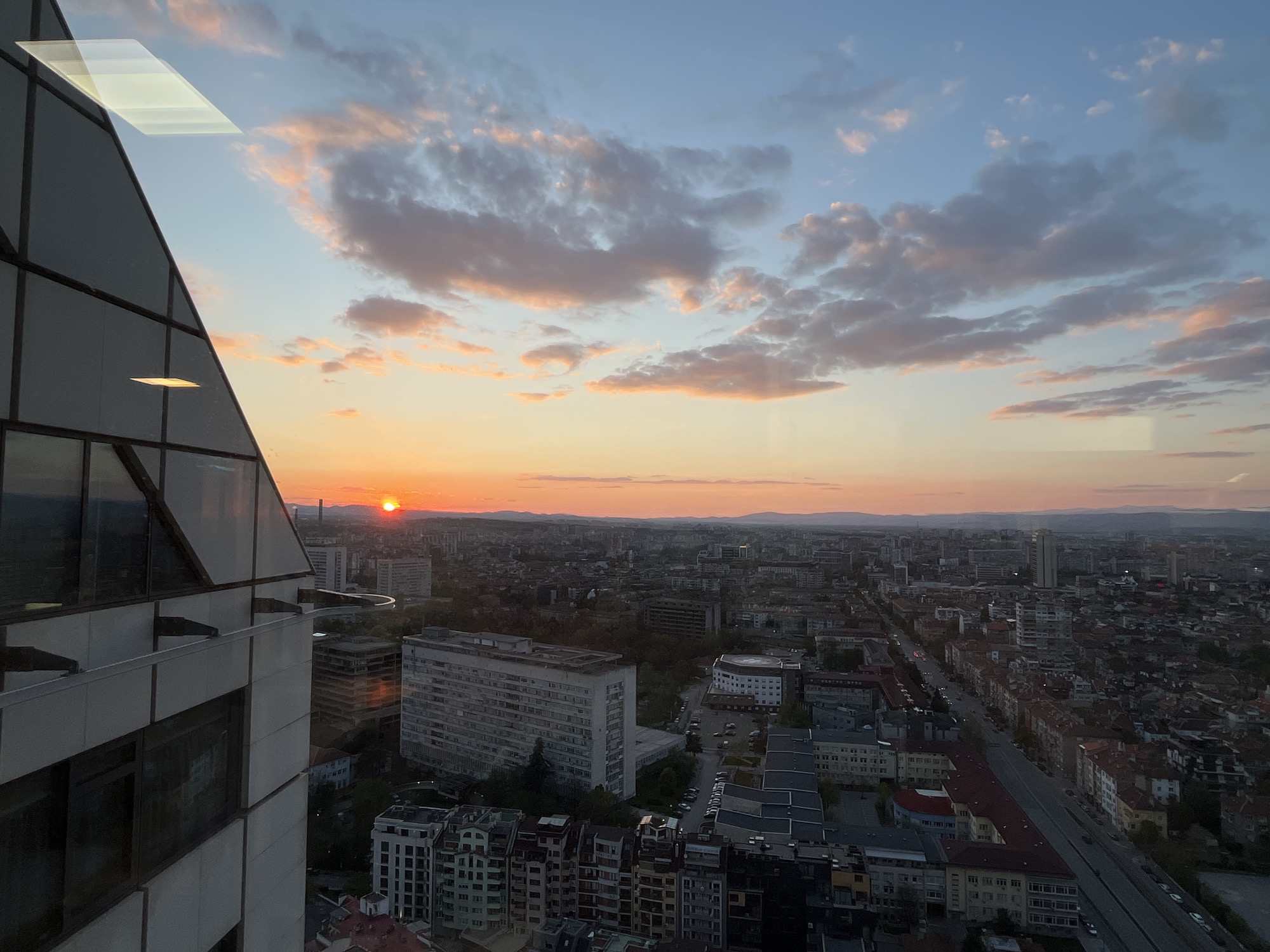 The image presents a wide panoramic view of Sofia, Bulgaria, captured during sunset from a high-rise building. On the left side, a section of the modern building from which the photograph is taken prominently features a glass and concrete facade, with angled panels reflecting ambient light.
The sky dominates the upper portion of the frame, showcasing a beautiful blend of colors as the sun sets. Near the horizon, the sky glows with warm golden-orange hues, transitioning upwards into a soft, clear blue. Scattered cumulus clouds, tinged with pink and orange by the low sun, are dispersed across the sky. The sun itself, a vibrant orange disc, is visible on the western horizon, slightly obscured by the distant landscape.
Below the colorful sky, the vast cityscape unfolds. A dense network of urban buildings, ranging from older residential blocks to more modern structures, stretches into the distance, appearing dark and silhouetted against the bright sky. Streets wind through the urban fabric, with some faint lights beginning to illuminate. In the far background, the subtle outlines of a mountain range or distant hills are visible against the horizon.
The overall scene captures a tranquil moment as the city transitions from day to night. There are no discernible human activities or specific events taking place, only the quiet grandeur of the urban landscape at dusk. No visible text is clearly legible on any of the buildings or objects within the frame.