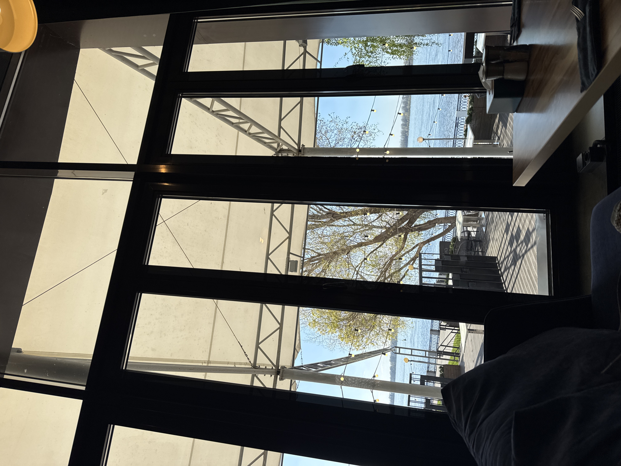 The image captures an interior view through a multi-paneled window, likely of a restaurant or cafe. Outside, a waterfront scene unfolds with a body of water visible, possibly a river or lake. Trees with sparse foliage, possibly indicating early spring, are strung with decorative lights. A boardwalk or patio area with tables and chairs is also seen. The interior is partially obscured by a dark cushion or seat back in the foreground, along with a table corner and a tissue box. The location is specified as the city of Unknown, Unknown.