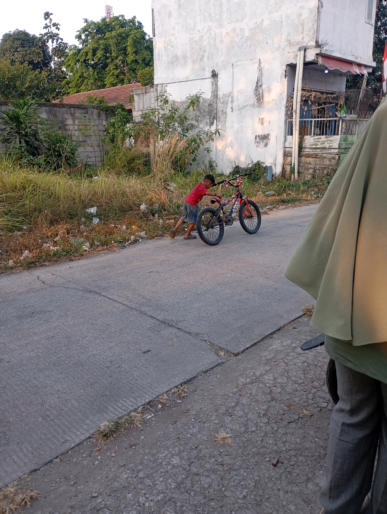 A young boy in a red shirt and blue shorts is pushing a red and black bicycle on an asphalt road in Babelan, Indonesia. He is walking on the right side of the road. The boy appears to be interacting with the bicycle, possibly preparing to ride it. The environment consists of a roadside with overgrown grass and debris, a concrete wall, and a building. A person in a light green garment is partially visible in the foreground, on the right side of the image, obscuring a portion of the road and the building. The scene takes place during daylight, indicated by the lighting and shadows.