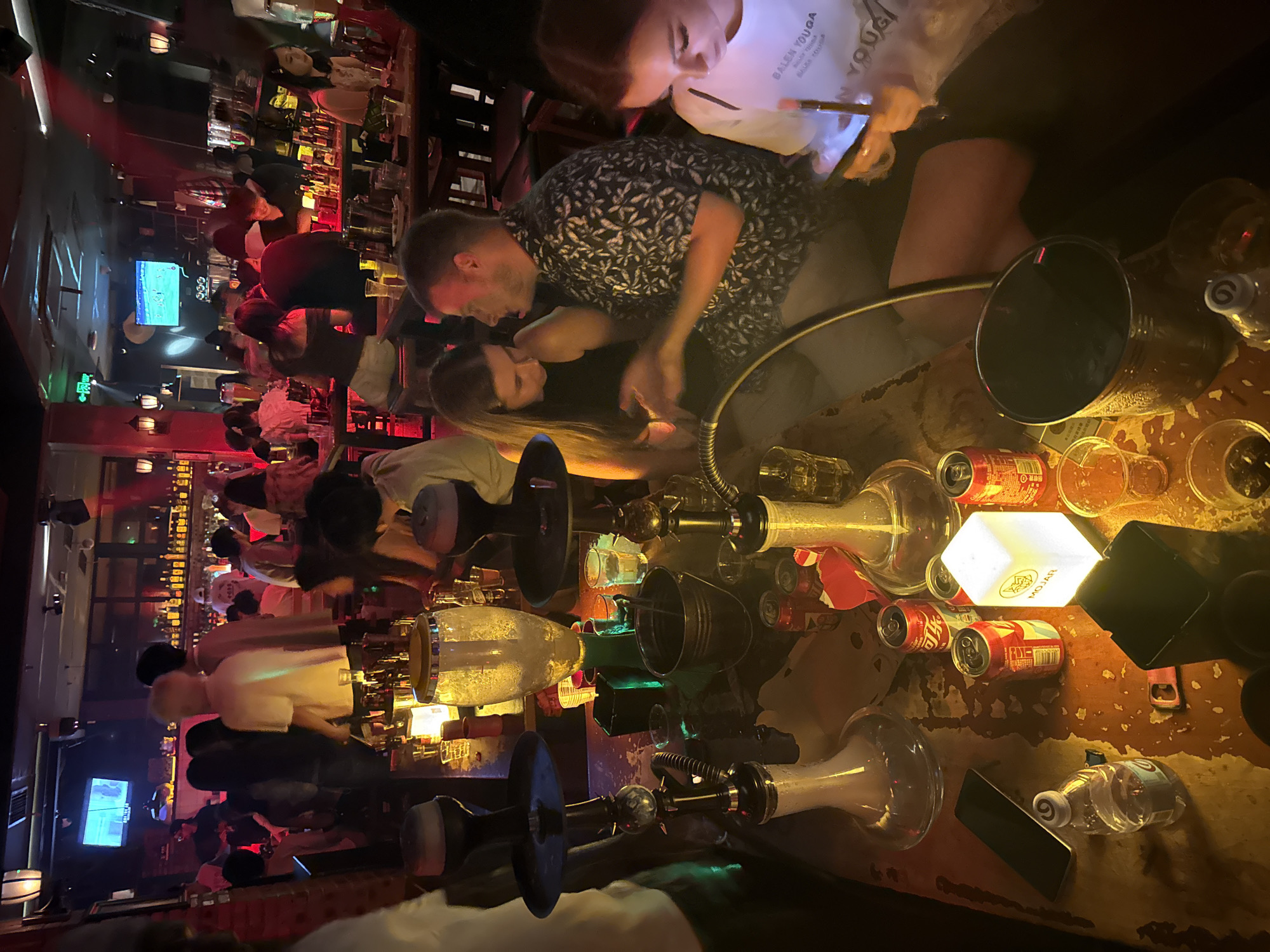 The image depicts a dimly lit interior of a bar or lounge, likely located in Liangjiang, China, given the provided context. The scene is filled with patrons engaged in various activities. In the foreground, on a wooden table, are two shisha pipes, several cans of what appear to be soft drinks, a bottle of water, a smartphone, a glowing cube-shaped lamp with the logo "MOJAR", and various cups.
Several individuals are visible at the table. A man and a woman are looking at a smartphone held by the woman, with the man gesturing. To their right, another woman is looking at her phone. Further back, a bar area is visible with shelves stocked with bottles and a television screen displaying some content. Other patrons are seated at tables in the background, some holding drinks. The overall atmosphere suggests a social gathering or night out.