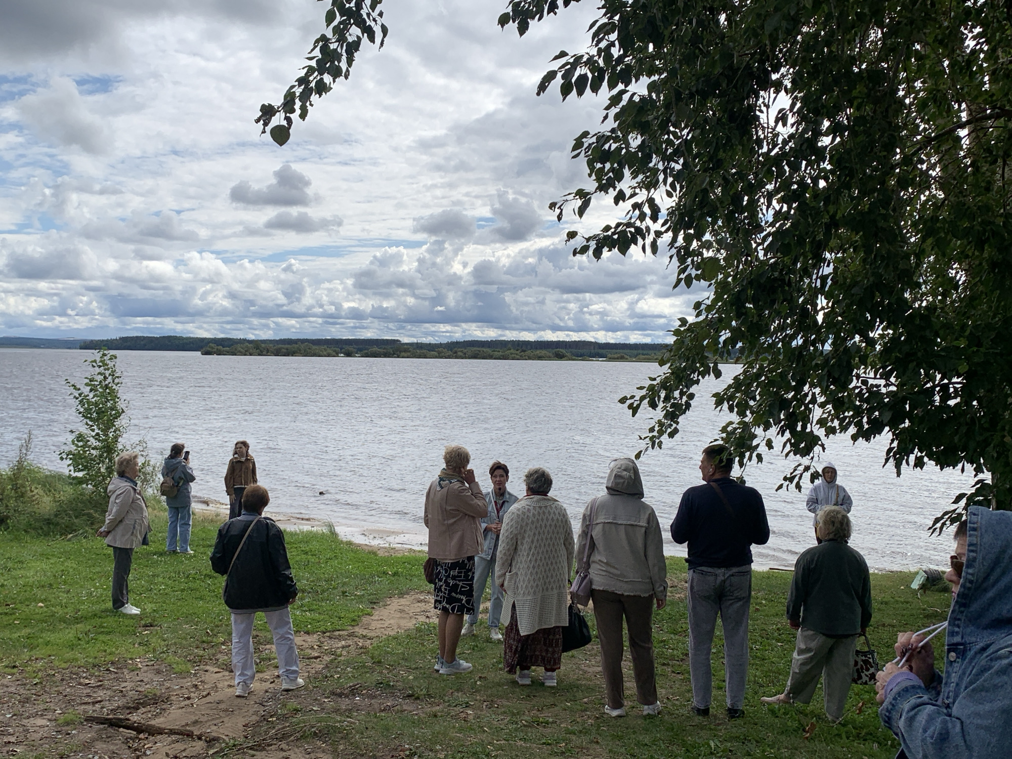 An outdoor scene depicts a group of people by a body of water under a cloudy sky. A wide river or lake stretches into the distance, bordered by a tree-lined horizon. Several individuals are scattered along the grassy bank. In the foreground, three women face away from the camera, one in a black jacket and light pants, another in a grey patterned sweater and dark skirt, and a third in a beige jacket. To the left, two women stand facing the water, one in a light-colored jacket and grey pants, and the other in a blue jacket and jeans, holding a phone to take a picture. Further in the background, a person in a light blue hooded jacket stands at the water's edge, and another person in a brown jacket is positioned near them. Two more individuals, one in a dark sweater and jeans, and another in a light grey hooded jacket, are also visible near the water. The environment is characterized by green grass, a sandy bank, and a large tree with dark green leaves partially obscuring the view on the right side of the frame. The sky is filled with a mix of white and grey clouds.