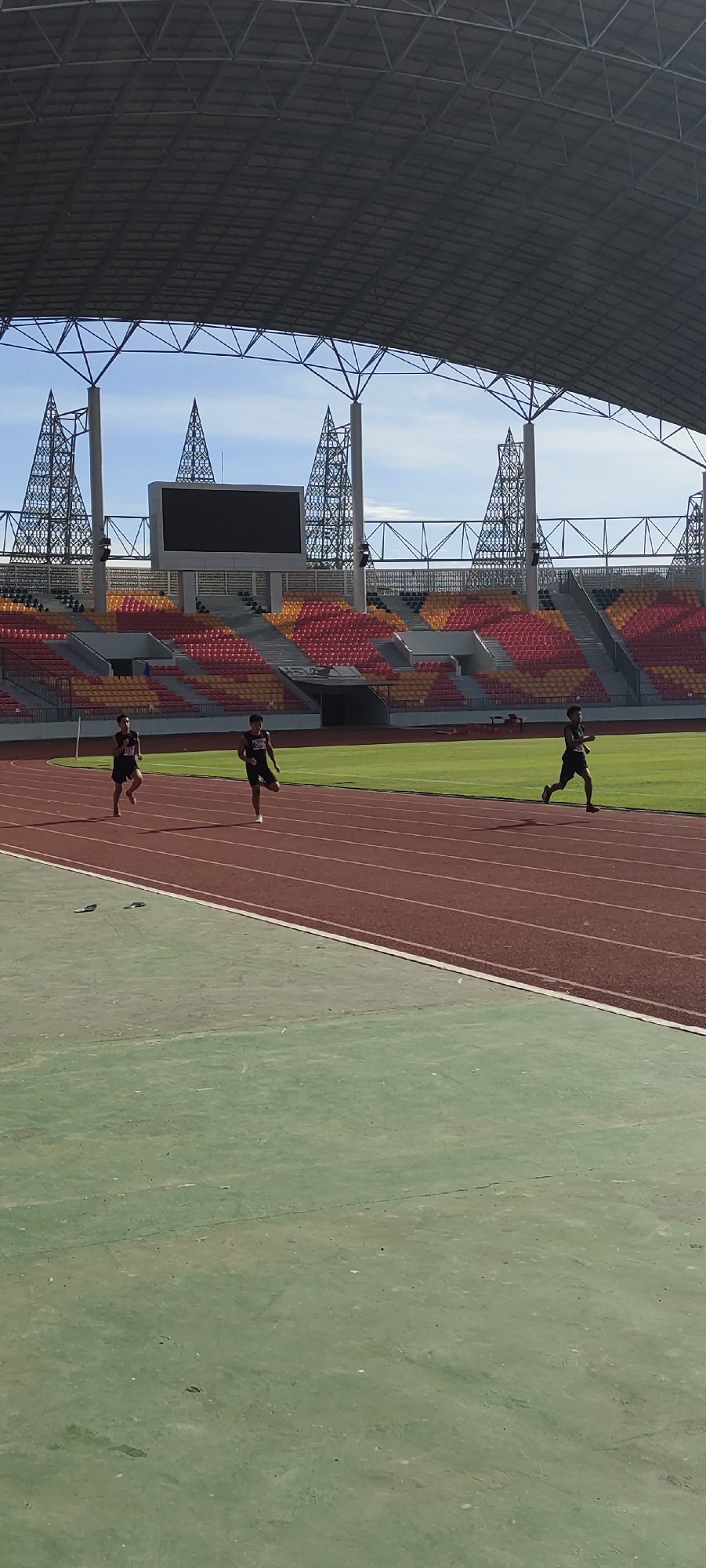 In Banda Aceh, Indonesia, three young men are running on a track within a stadium. They are wearing dark athletic shorts and t-shirts, some with visible bib numbers. The track is made of red synthetic material, with white lane markings. In the background, a large stadium with tiered seating in shades of red and yellow is visible, along with a large Jumbotron screen. The stadium is covered by a large, arched roof supported by metal structures. The sky is blue with some scattered clouds. The foreground shows a green area, likely a field or another part of the stadium's infrastructure. The scene appears to be a sports training or competition event.