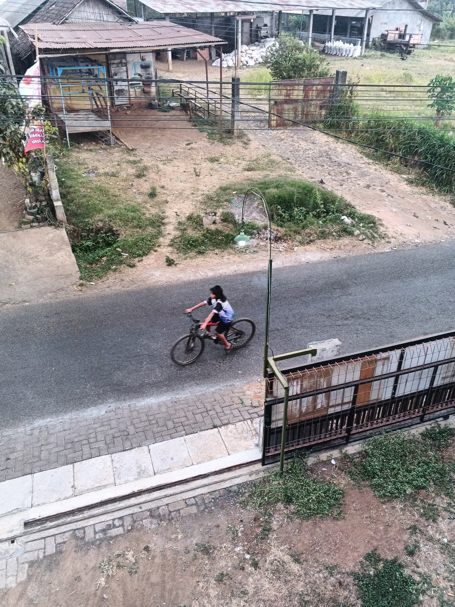 The image, captured from an elevated perspective, depicts a street scene in the city of Bocek, Indonesia. A young person is riding a bicycle on the asphalt road, which runs parallel to a brick-patterned sidewalk and a dirt area with sparse vegetation. To the right of the road, a metal fence with rectangular panels separates the street from an area with overgrown grass and a lamp post. Beyond the fence, buildings with corrugated metal roofs are visible, along with a large, open structure. Further in the background, a field with tall grass stretches towards other structures, including what appears to be a building with stacked white bags and a trailer. The overall environment suggests a rural or semi-rural setting within Bocek.