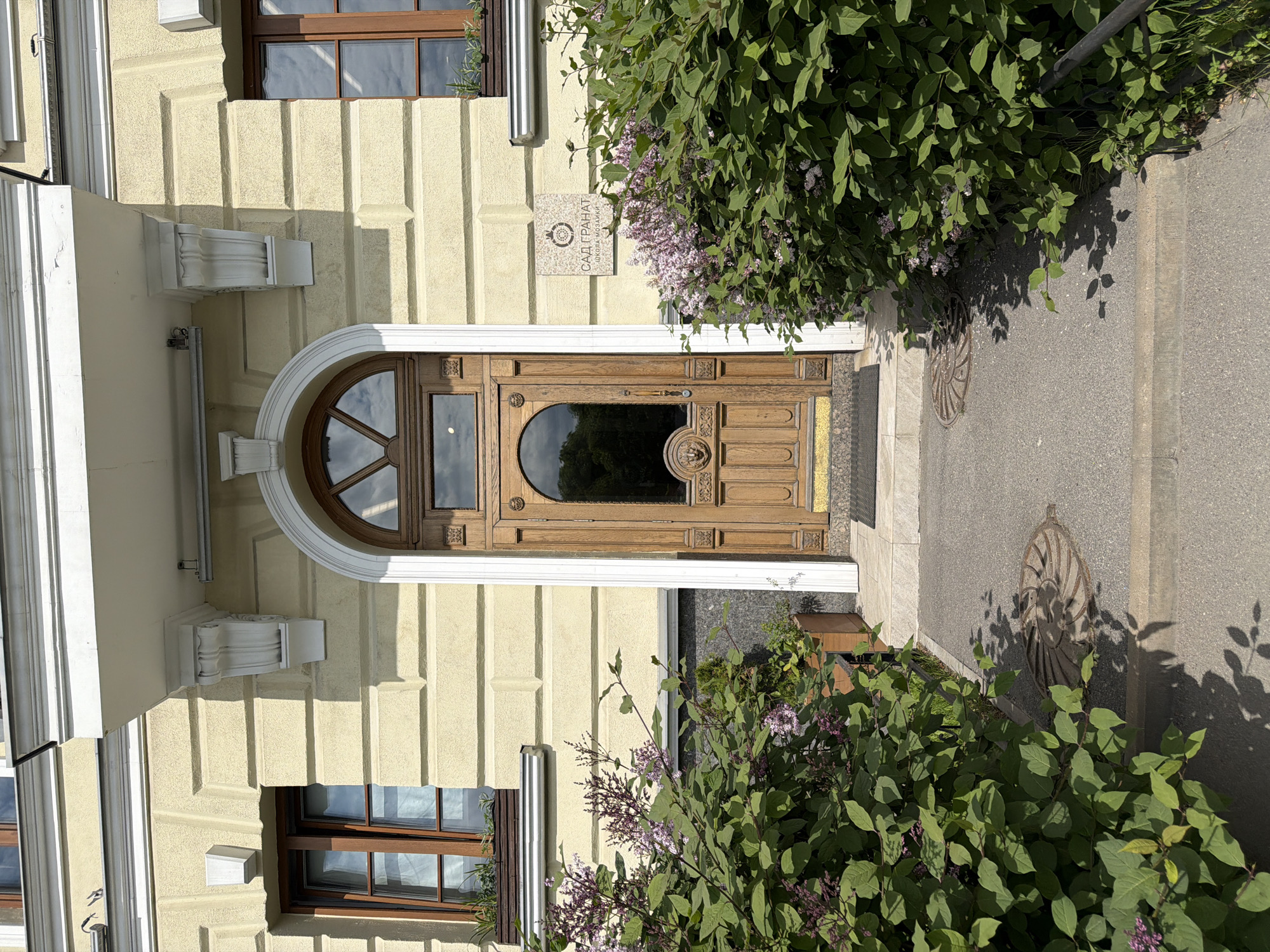 This image, taken in Saint Petersburg, Russia, displays the entrance of a building on a sunny day. The building features a light cream-colored facade with vertical fluting patterns and white decorative architectural elements, including ornate brackets. Dark-framed windows are visible on either side of the main entrance.
The central focus is a grand, carved wooden door set within a prominent white arched entryway. The door is intricately designed with detailed panels and a large, rounded arched glass pane in its upper section, reflecting the sky.
To the right of the door, a square mosaic plaque is mounted on the wall, partially obscured by foliage. The Cyrillic text on the plaque reads: "САД ГРАНАТ" (SAD GRANAT), which translates to "Pomegranate Garden," and below it, "ШКОЛА МОЗАИКИ" (SHKOLA MOZAIKI), meaning "School of Mosaics."
Lush green bushes with small clusters of purple flowers, likely lilac, flank both sides of the entrance, casting strong shadows on the facade and the pavement. The pavement itself is light grey and features an embedded circular metal grate with a distinctive spiral design on the right side. The clear skies and sharp shadows indicate a bright, sunny day.