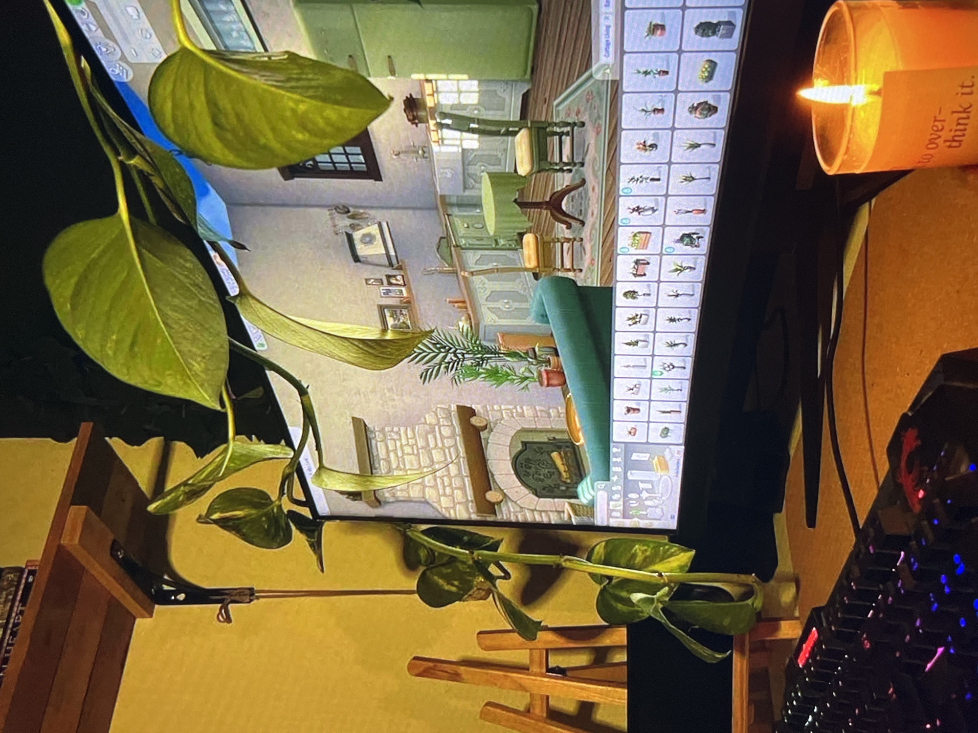 The image depicts a computer monitor displaying The Sims 4. The game is in build mode, showing a room with a fireplace, furniture, and build assets visible in the right panel. A live candle with the text "to over-think it" is situated to the right of the monitor. A computer keyboard with illuminated keys is visible in the lower right corner. A houseplant is draped across the left side of the monitor, obscuring part of the screen. The setup is located in an interior space.