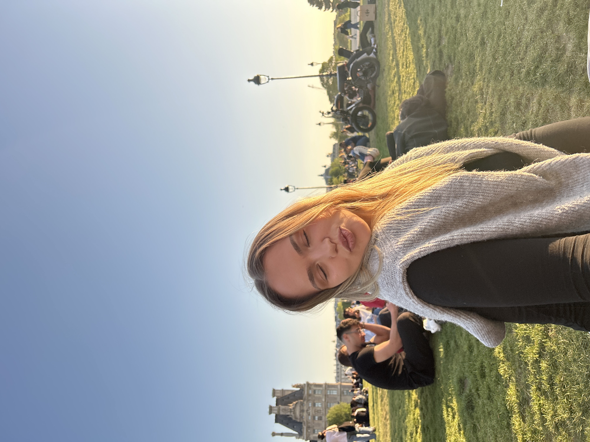A selfie features a young woman with blonde hair, eyes closed, and lips slightly pursed in a playful expression. She is wearing a light grey, possibly knitted, sleeveless top over dark long-sleeved clothing. Golden sunlight warmly illuminates her hair and face from the right side of the frame.
The outdoor scene depicts a large, well-maintained green lawn, likely a park, under a clear blue sky. The lighting suggests it is late afternoon or early evening, with soft, warm sunlight casting gentle shadows on the grass. In the background, numerous people are scattered across the lawn, sitting and lounging, indicating a leisurely gathering or public relaxation. Several motorcycles or scooters are parked in the mid-ground, alongside classic black lampposts. Further in the distance, a grand, ornate building with intricate architectural details, typical of a historical palace or chateau, is visible.
The overall setting points to a public park in Paris, France, where individuals are enjoying the sunny weather and open space. No visible text is present in the image.