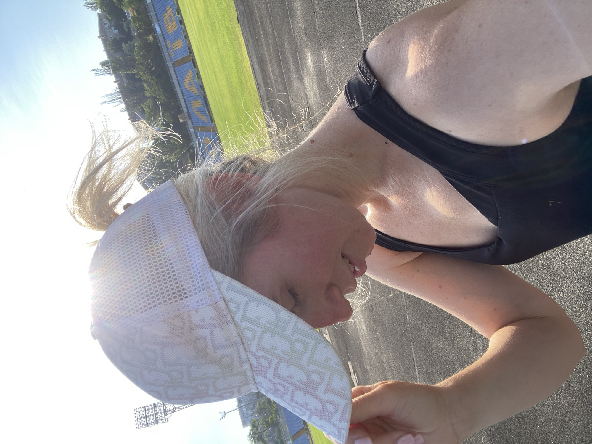 A woman is outdoors at a stadium, wearing a white trucker hat and a black tank top. Her hair is pulled back into a ponytail and some strands are blowing in the wind. The woman is looking down and to her right, holding the brim of her hat with her left hand. The stadium has a track and field and blue and yellow bleachers in the background, with buildings and trees visible on a hill behind the stadium. The sun is shining brightly, casting a warm glow on the scene.