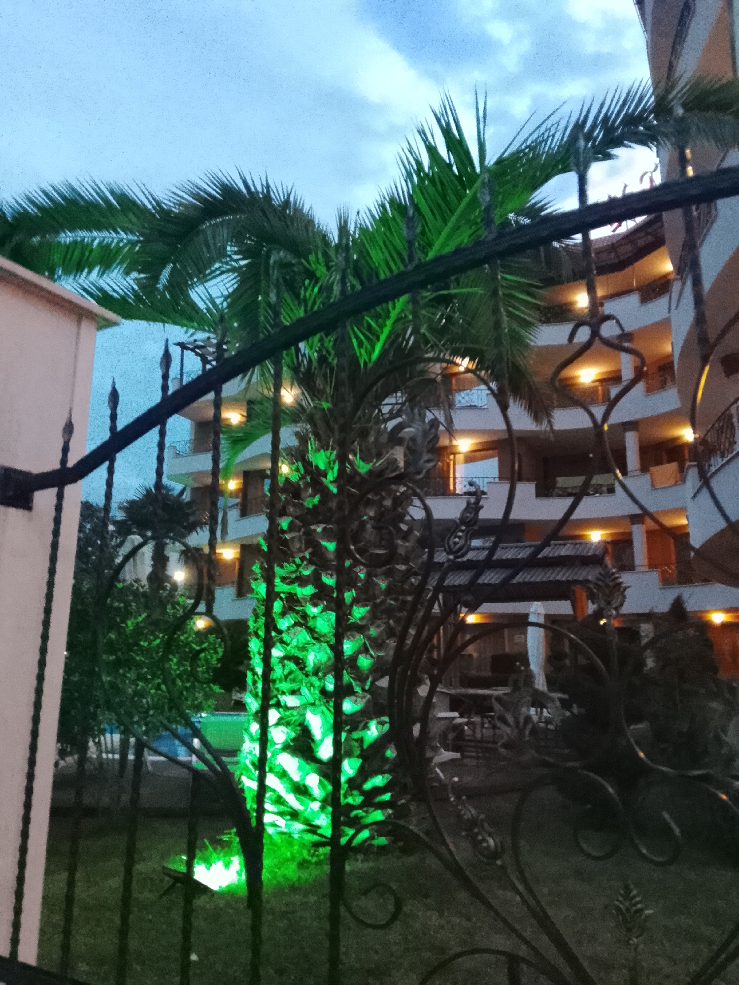 The image shows a palm tree illuminated with green lights at dusk, in front of a multi-story building with balconies. The building appears to be a hotel or apartment complex, with lights visible in some of the windows. A black metal fence is in the foreground, obscuring part of the scene. The sky is cloudy and appears to be a twilight blue. The scene is likely a hotel courtyard or garden area in Nesebar, Bulgaria, given the architectural style and vegetation. There are no people visible in the image.