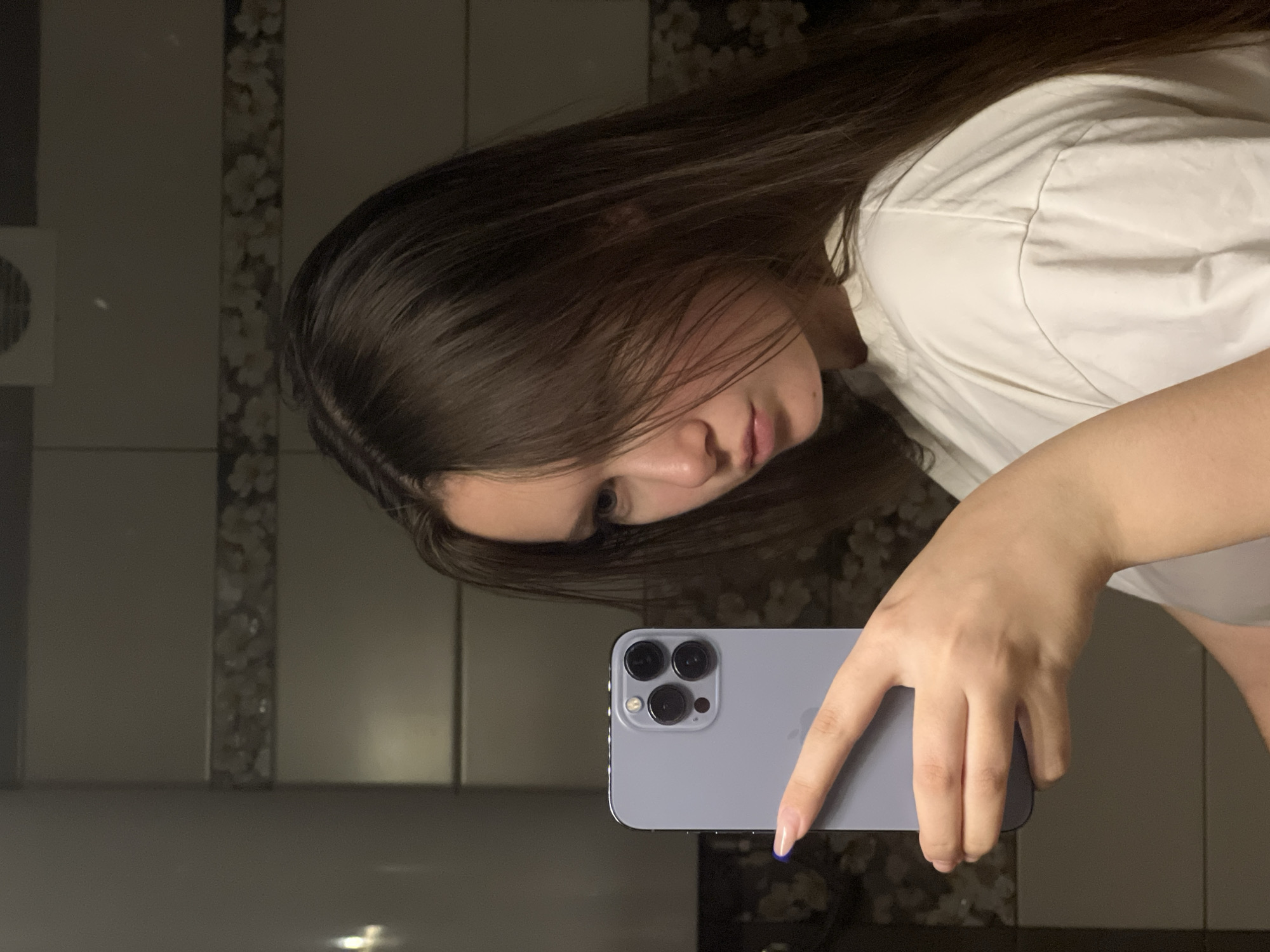 A young woman with long, dark brown hair is taking a selfie in what appears to be a bathroom. She is holding a light blue iPhone with three camera lenses. She is wearing a white shirt. The background consists of light gray tiles with a decorative strip of floral pattern. The scene is likely in an indoor location, potentially the city of Chaikovsky, Russia.