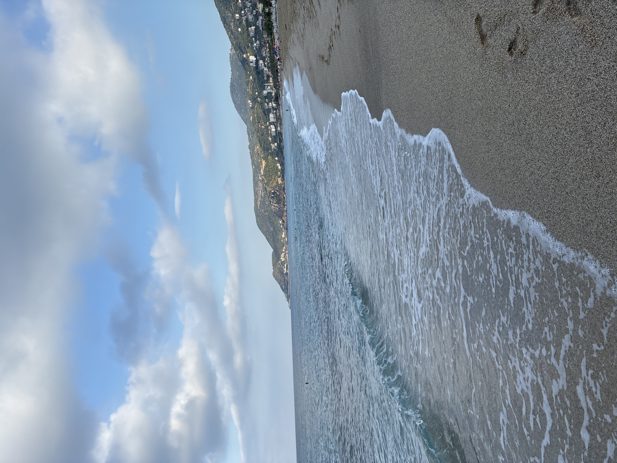 A wide, sandy beach is bordered by a calm, blue sea with gentle waves. On the left side, the water is a clear blue, with a few ripples and foam from the incoming tide. The sand is a light brown and has footprints. In the distance, a lush green hillside dotted with white buildings and trees slopes down towards the sea. The sky above is a mix of blue and scattered white clouds. The scene is a tranquil coastal landscape.