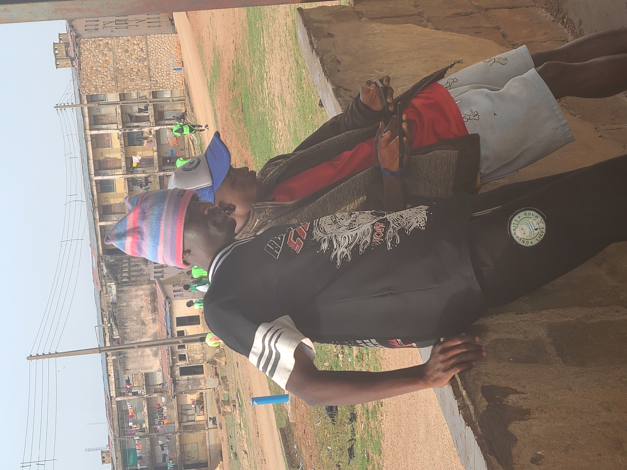 Two men are seated, one with a striped beanie and the other with a blue and white baseball cap, both appearing to be looking at mobile phones. They are positioned against a concrete structure. The background reveals a cityscape with aged buildings, laundry lines, and an open field. In the distance, several figures in green vests are observed. The location is likely Jos, Nigeria.