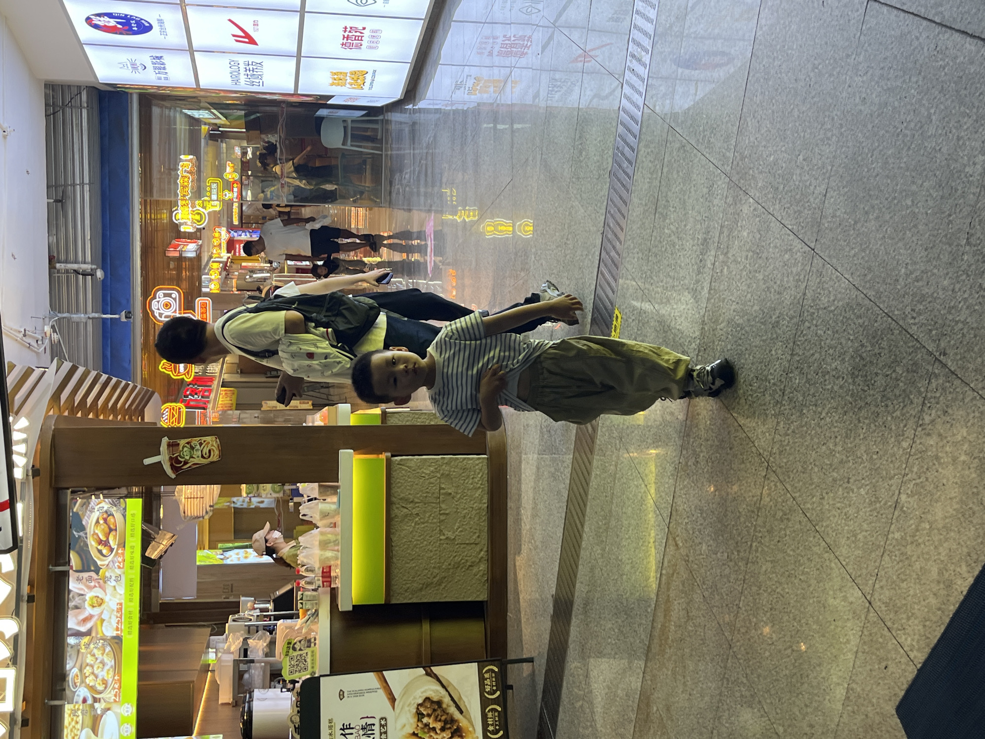 A young boy in a striped t-shirt and loose pants walks towards the camera in an indoor marketplace. He is in the foreground, slightly off-center to the left. Behind him, a woman in a white t-shirt and black pants walks away from the camera, carrying a backpack. The marketplace has bright lighting from overhead fluorescent fixtures and neon signs. On the left, a food stall displays images of dumplings and has a woman behind the counter. On the right, a wall features illuminated panels with various logos and Chinese text, including "HAROLOGY" and "德香苑". The floor is tiled with a reflective surface. The scene appears to be in Yuzhong District, China.