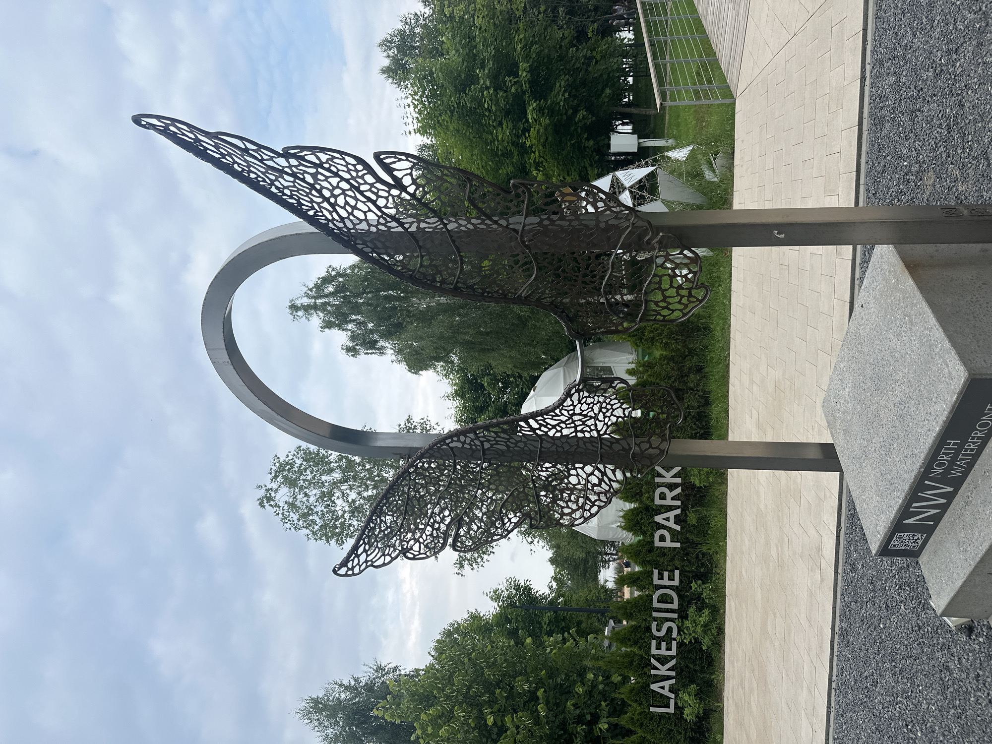The image shows a metal sculpture of wings in a park setting. The sculpture is positioned in front of a sign that reads "LAKESIDE PARK" and a row of evergreen trees. Behind the trees, there are some geodesic domes. The ground in the foreground is covered with dark gravel, and there is a paved walkway. To the right, a wooden walkway with railings is visible. The sky is overcast. The sculpture itself is made of dark metal with an intricate, lace-like pattern. It is supported by a metallic post. At the base of the sculpture, there is a stone structure with a plaque that includes the letters "NW" and the words "NORTH WATERFRONT" and a QR code. The overall impression is of a modern public space with an artistic focal point. This sculpture is located in the North Waterfront Park in Minsk, Belarus.