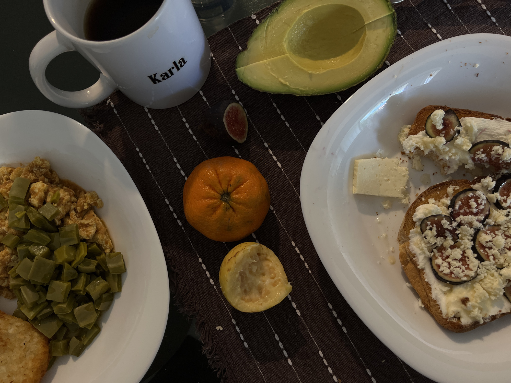 The image shows a tabletop setting with various breakfast items. On the left, a white plate contains scrambled eggs with nopales (cactus pads) and a hash brown. A white mug with the name "Karla" in black lettering contains a dark liquid, possibly coffee or tea. To the right, another white plate features two pieces of toast topped with cream cheese and sliced figs, accompanied by a piece of white cheese. Additional food items are scattered on the surface, including half an avocado, a sliced fig, an orange, and a partially eaten guava. The scene is set on a brown placemat with a white striped pattern. The overall environment suggests a home or restaurant setting. There is no evidence to confirm the location is San Bernardino Tlaxcalancingo, Mexico.