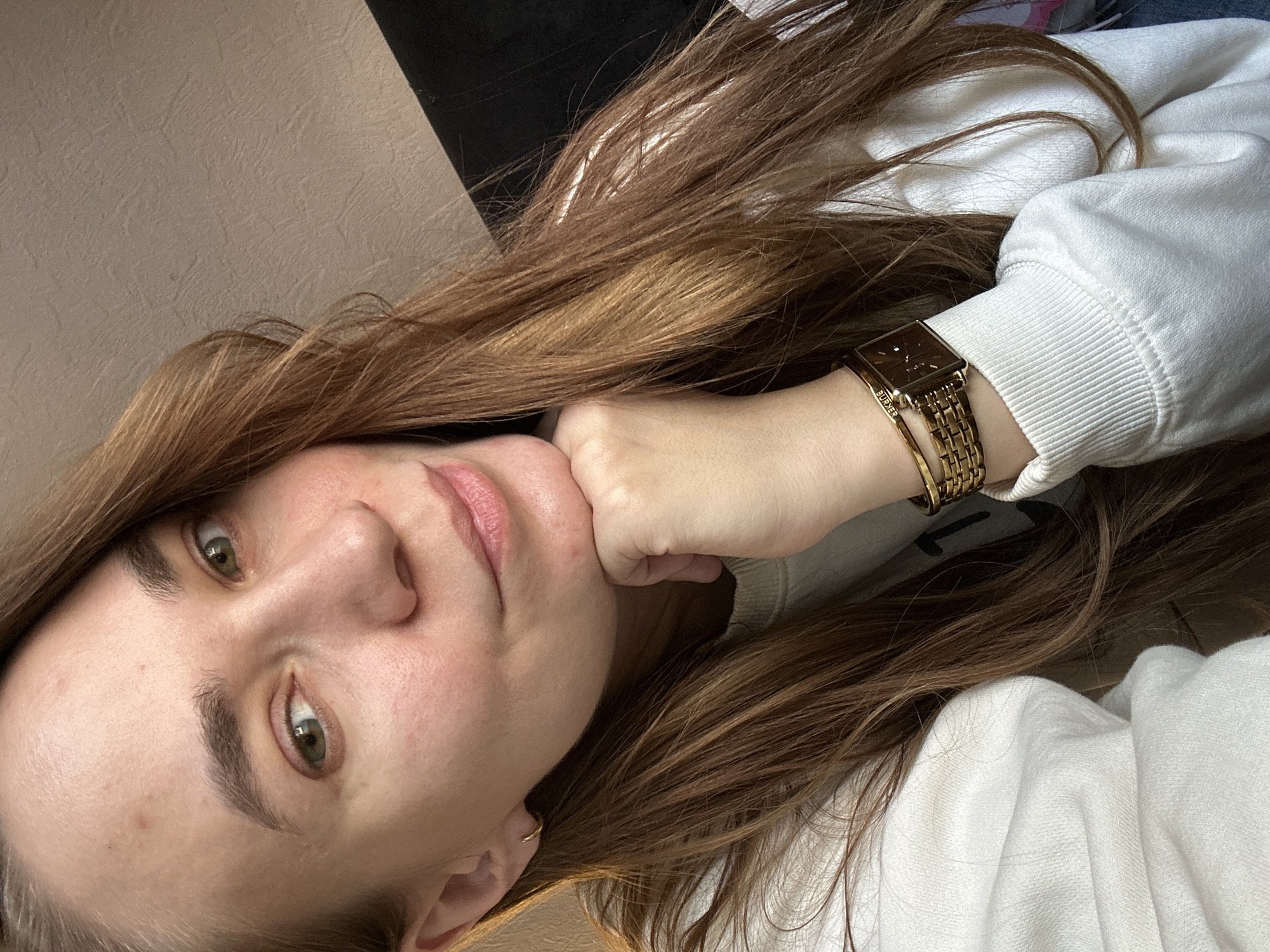A close-up selfie-style shot of a woman with light brown hair, light skin, and green eyes. She is wearing a white sweatshirt and a gold watch on her left wrist. Her left hand is resting under her chin. The background includes a light beige wall and a dark-colored object, potentially furniture, at the top. There is no indication of location within Chaikovsky, Russia, or any other city.