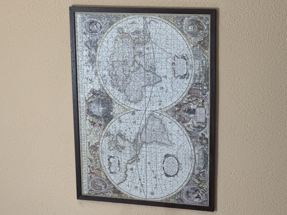 A framed jigsaw puzzle of an antique world map is mounted on a light beige, textured wall. The map, titled "NOVA TOTIUS TERRARUM ORBIS GEOGRAPHICA AC HYDROGRAPHICA TABULA" and attributed to "Auth. Henr. Hondio," displays two hemispheres, with the Americas on the left and Eurasia and Africa on the right. Intricate decorative borders feature figures, cartouches, and navigational elements. The puzzle is encased in a dark, simple rectangular frame. No individuals, other objects, or specific room identifiers are visible. The exact location within Blagoevgrad, Bulgaria, cannot be determined from the image.