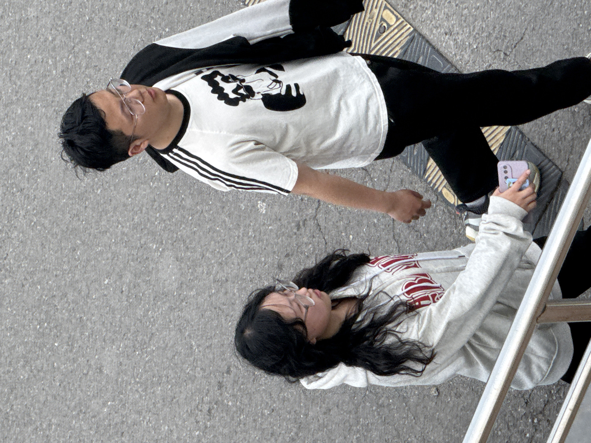 Taken from an overhead perspective, this image captures two individuals, a male and a female, on a concrete outdoor surface in Zhongtun, China. The lighting is soft and diffused, indicating a cloudy day or a shaded environment.
The male subject is positioned diagonally across the frame. He wears a white short-sleeved t-shirt with black stripes on the sleeves, likely from Adidas, and a black graphic design of a stylized character or face on its chest. He also has black pants and clear-framed glasses. His dark hair is neatly styled. A section of yellow tactile paving with raised lines is visible beneath his feet, suggesting a public walkway or a path for the visually impaired.
The female subject is located towards the bottom right, next to a metal railing. She has long, dark, wavy hair and wears clear-framed glasses. Her attire includes a light grey hooded sweatshirt with a red, stylized graphic design on the front, and black pants. She is actively engaged with a light purple smartphone, holding it in her hands and looking at its screen. A small, lighter-colored design is visible on her phone case.
The ground is a textured grey concrete. The metal railing suggests a barrier, staircase, or other urban infrastructure element. The scene depicts a casual moment in a public setting.