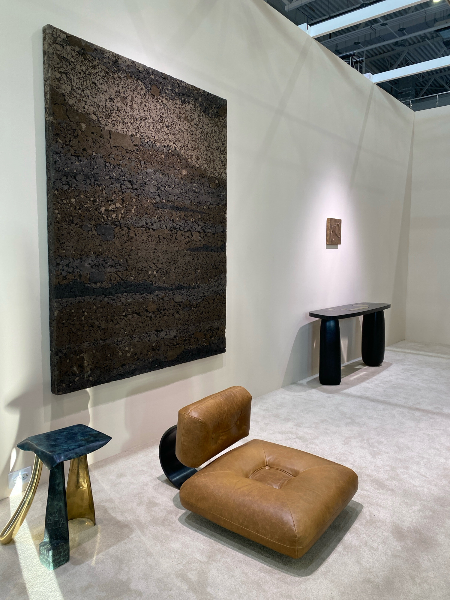 An interior scene, likely an art exhibition or gallery, displays contemporary furniture and artwork. In the foreground, a brown leather armchair with a black curved base faces a small, dark green and brass side table with an irregular top and three uniquely shaped legs. Behind the armchair, a long, dark console table with rounded legs supports a small, rectangular bronze artwork mounted on the wall. To the left, a large, textured dark brown artwork hangs on a white wall. The floor is covered with a light-colored carpet. The scene is lit by overhead track lighting and a spotlight illuminating the console table artwork. The overall impression is one of minimalist design and artistic presentation, with a focus on textured materials and sculptural forms. The location is stated to be in the city of Khimki, Russia. There are no people visible in the image.