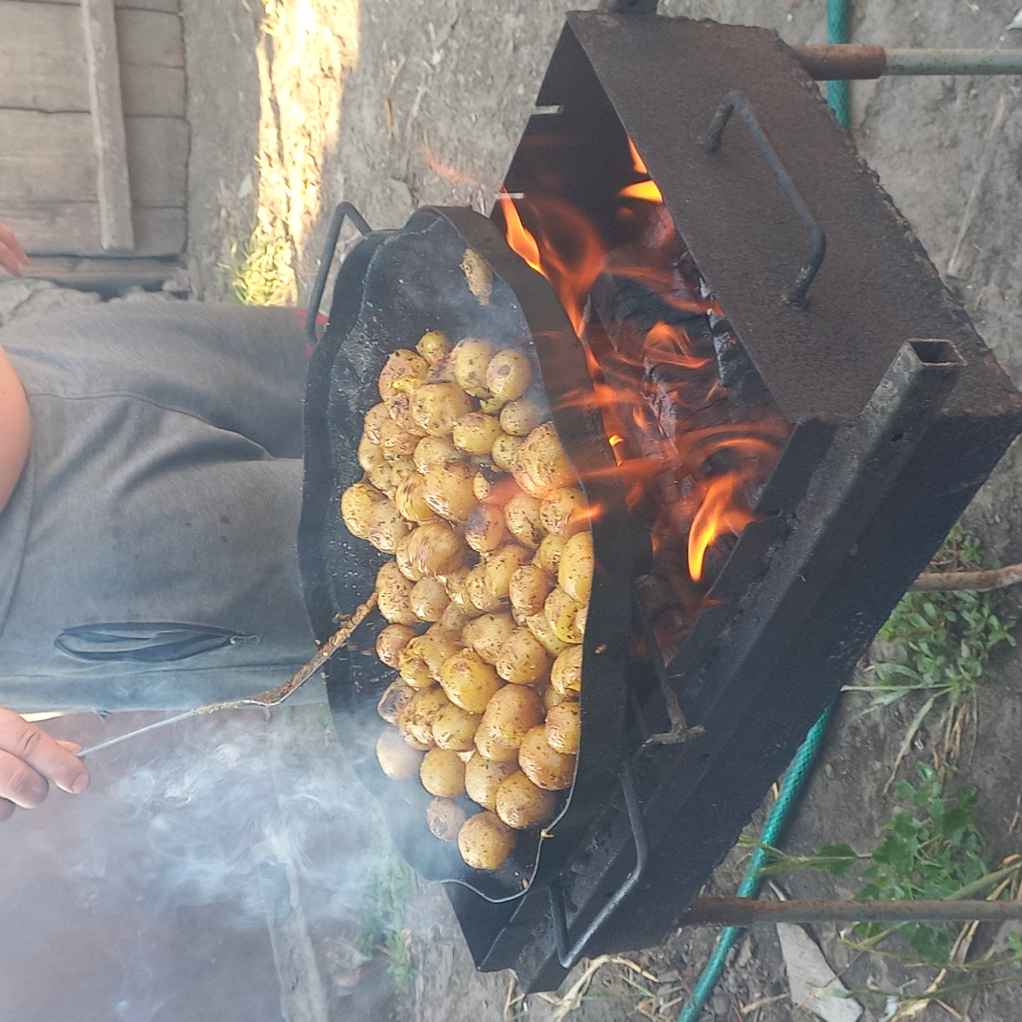 A close-up shot captures a person's hand holding a metal spatula, stirring a large quantity of small, yellow potatoes in a black, round, shallow pan. The pan is elevated over an open-air grill filled with glowing embers and active flames. Wisps of smoke rise from the pan, obscuring the background slightly. The potatoes appear to be seasoned with visible specks of herbs or spices and are showing signs of browning from the cooking process. The person is wearing grey sweatpants, partially visible in the upper portion of the frame. The surrounding environment suggests an outdoor setting with dry ground, scattered debris, and some green foliage in the lower left. The grill itself is dark, metallic, and appears weathered. The scene is lit by natural sunlight, creating highlights and shadows. The image does not provide any specific visual cues to indicate the location as Klyntsi, Ukraine.