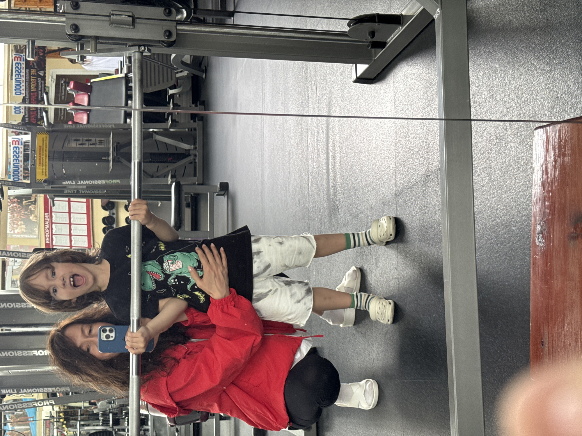 An adult woman and a young child are captured in a mirror selfie within what appears to be a gym or fitness center in Chaikovsky, Russia. The woman, with long brown hair, is in the foreground, wearing a red jacket over a dark top and black pants. She is holding a blue smartphone to take the picture, partially obscuring her face. Her left hand rests gently on the child's back.
The child, likely a boy with long light brown hair, stands behind and slightly to the left of the woman. He wears a black t-shirt with a green dinosaur graphic, white tie-dye shorts, and light-colored clogs paired with striped socks. He has an excited, open-mouthed expression, looking towards the camera.
A metallic horizontal bar, part of the gym equipment, runs across the mid-ground of the image. The background reflects more gym equipment, including red-padded benches and various metal structures, indicating an indoor fitness setting. The floor is dark grey.
Visible Cyrillic text on reflected signs in the background includes "ПРОФЕССИОНАЛЬНАЯ ЛИНИЯ" (PROFESSIONAL LINE) and "ИНФОРМАЦИЯ" (INFORMATION). Additionally, some reversed Latin characters and numbers like "EZZEU010" are discernable. The scene suggests a lighthearted, playful moment shared between the two individuals.