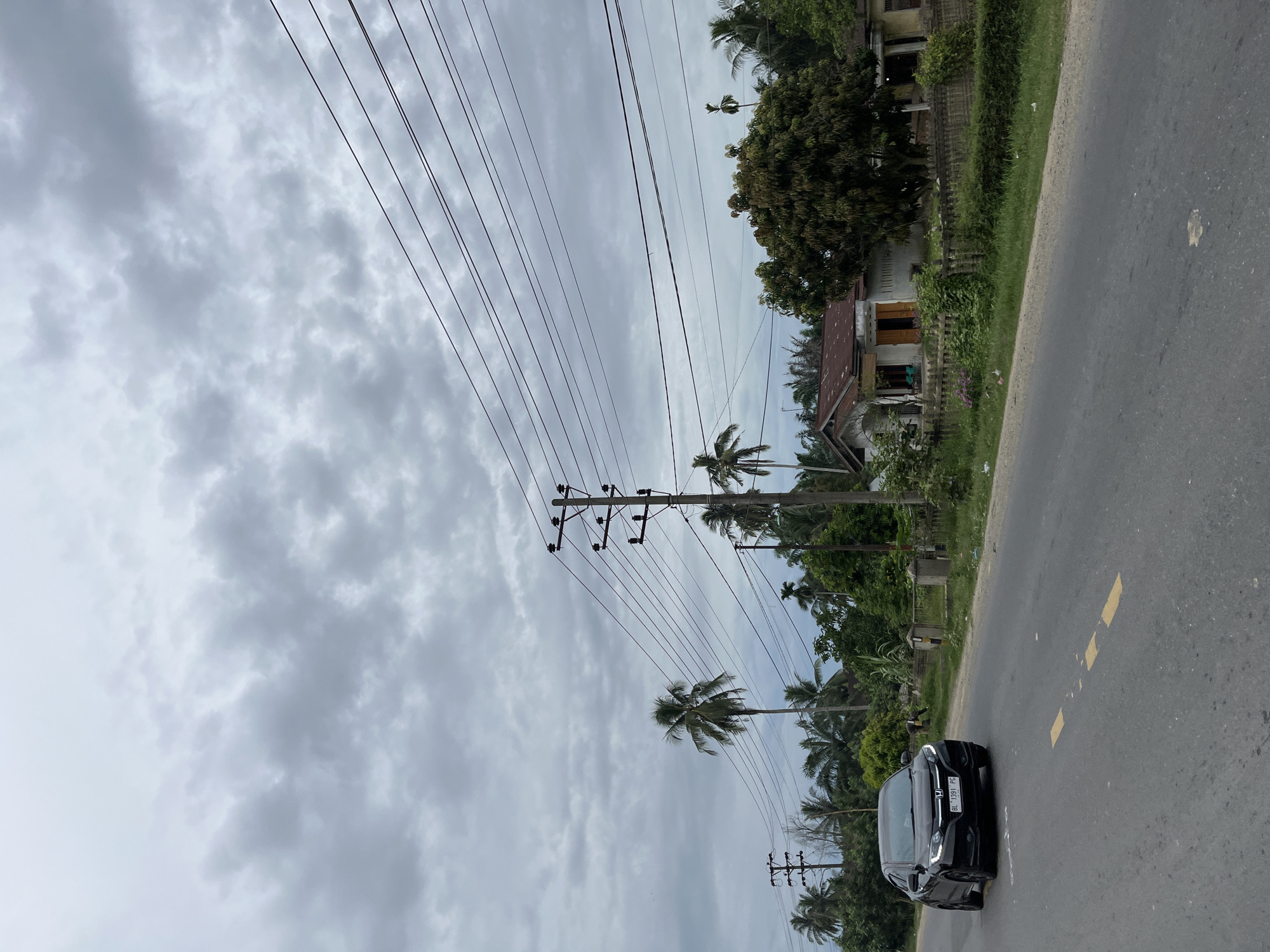 The image shows a road in Simpangempat, Indonesia, with a black sedan driving on the left side of the asphalt road. The car has a license plate that reads "BL 1391 PG". On the right side of the road, there is a house with a reddish-brown roof and several trees surrounding it. Several utility poles with power lines run across the scene, with a cloudy sky overhead. The vegetation is lush and green, with palm trees visible in the background.