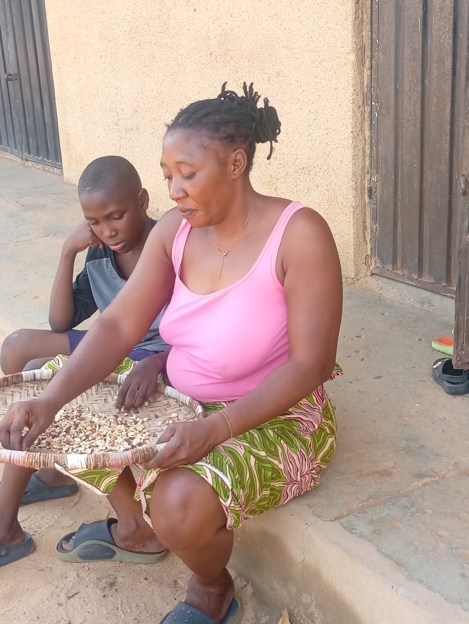 A woman and a boy are sitting on a concrete step outside a building in Yelwa, Nigeria. The woman, wearing a pink tank top and a green and pink patterned skirt, is sorting beans in a flat woven tray. The boy, wearing a dark shirt and shorts, sits beside her, his head resting on his hand. The building has a beige wall and a dark metal door. The ground is dusty. A pair of sandals sits on the step near the door.