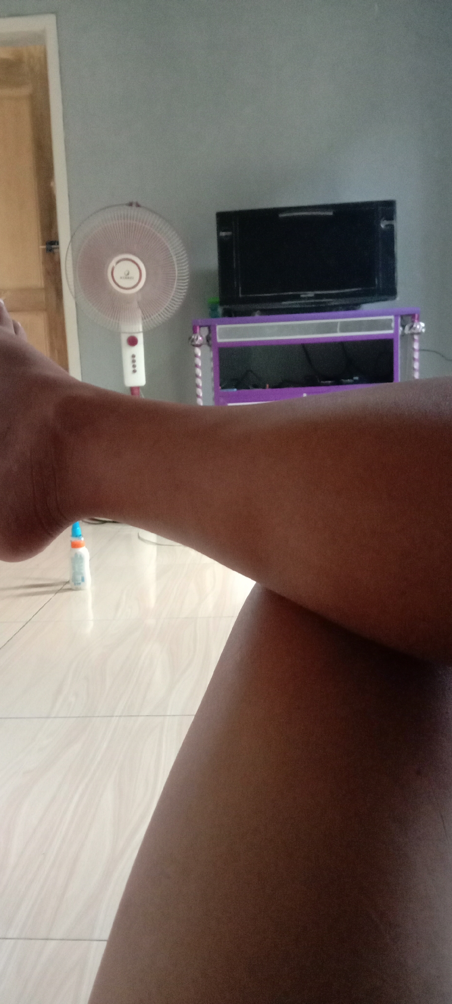 An indoor, low-angle, medium shot shows a person's legs crossed and resting on a tiled floor. In the background, a white oscillating fan stands to the left of a purple media stand topped with a black television. The room appears to be in a residential setting in Utama Jaya Mataram, Indonesia.