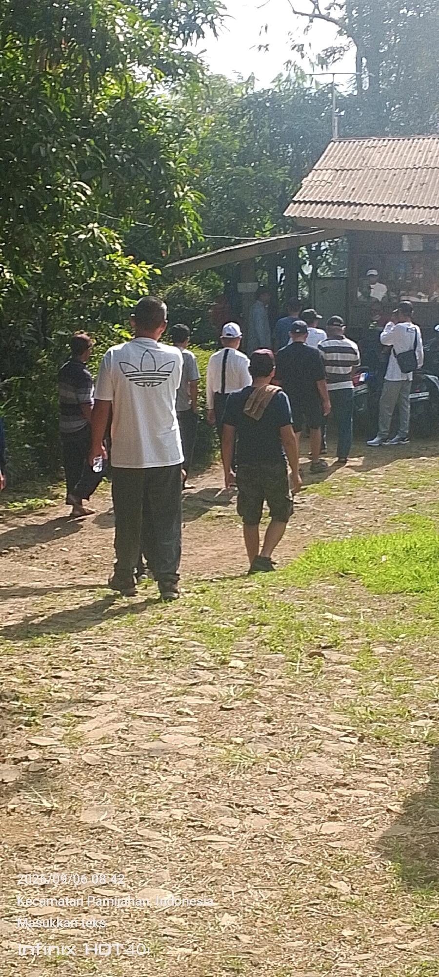 In Pamijahan, Indonesia, a group of men are walking on a dirt path toward a small, open-air building with a corrugated metal roof. The building is partially obscured by trees and foliage. The path is uneven, with scattered rocks and patches of grass. Several men are walking away from the camera, while one man in the foreground faces away from the camera. He is wearing a white t-shirt with a black Adidas logo and dark pants. Another man to his left is wearing a striped shirt and dark pants, and is holding a bottle of water. The overall atmosphere appears to be casual and outdoor.