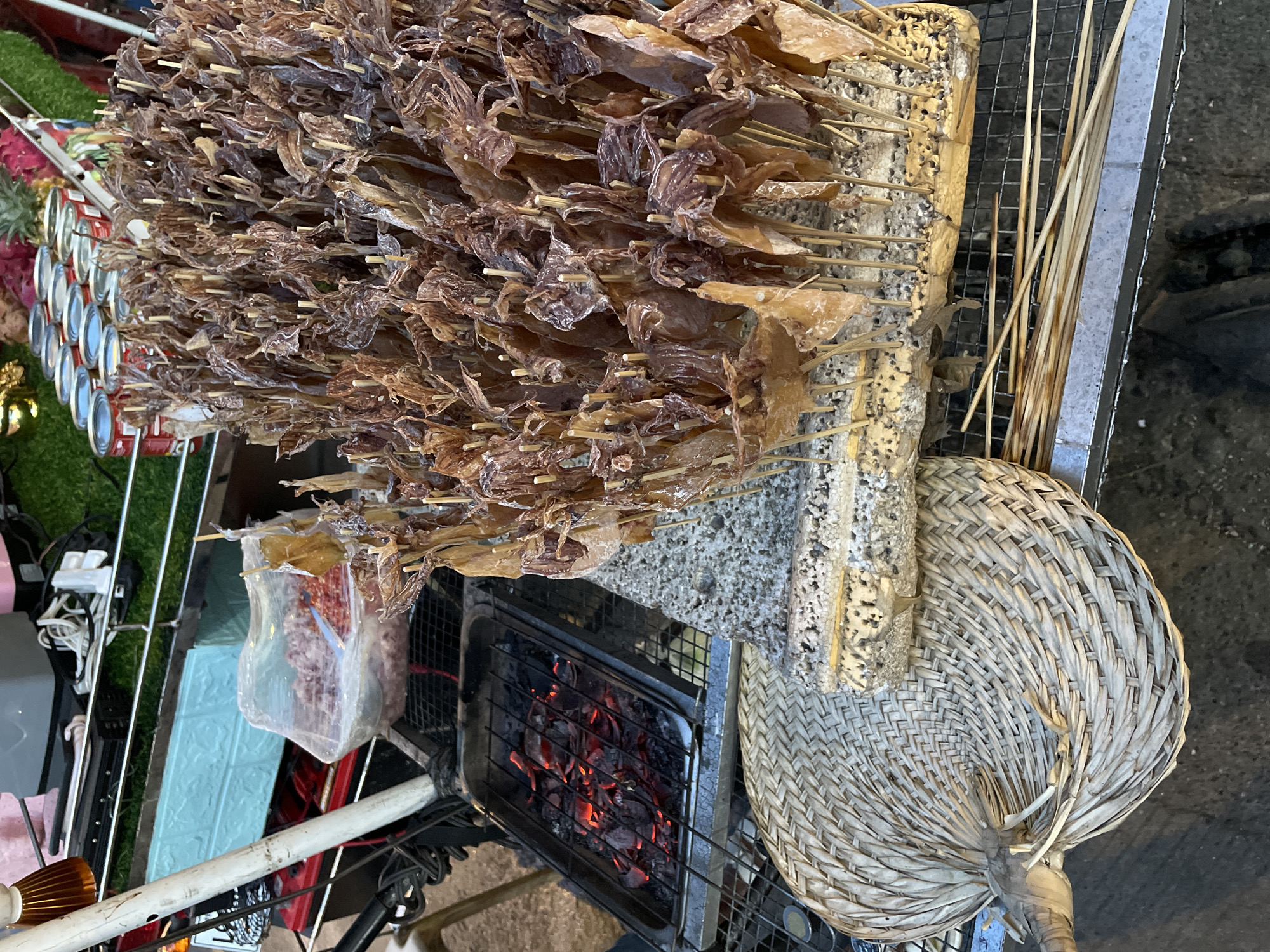 This image depicts a street food stall in Davao City, Philippines, specializing in grilled squid. The stall features a variety of items related to food preparation and display. In the foreground, a large, woven fan lies on a metal grate. Behind the fan, a charcoal grill with glowing embers is visible in a metal tray, indicating active cooking.
The main subject of the image is a large display of dried squid skewers, standing upright on a wooden base. The squid are skewered on thin wooden sticks and appear to be prepared for grilling. To the left of the squid display, a plastic bag filled with ground meat, possibly for other dishes, is placed on a shelf. In the background, a rack holds several cans, and a section of green artificial turf is visible, suggesting a decorated stall. The overall impression is a bustling street food environment.