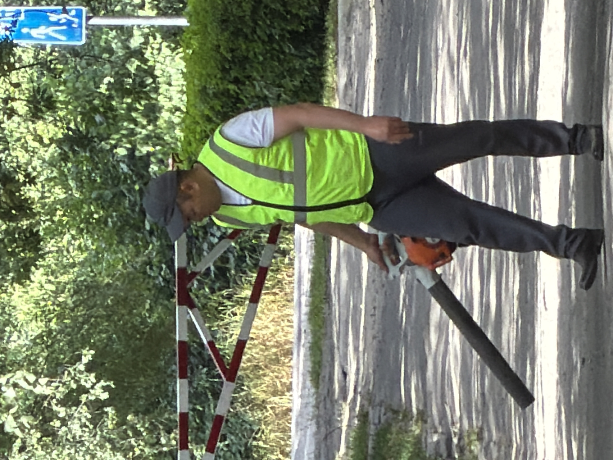 A man wearing a high-visibility lime green vest over a white t-shirt and dark pants operates a leaf blower. He is wearing a dark baseball cap and black shoes. The man is positioned on a paved surface, possibly a road or pathway, with a red and white striped barrier to his left. Behind him is lush green foliage and trees. To the right of the image, a blue sign with white figures of people is visible on a pole. The lighting suggests it is daytime.