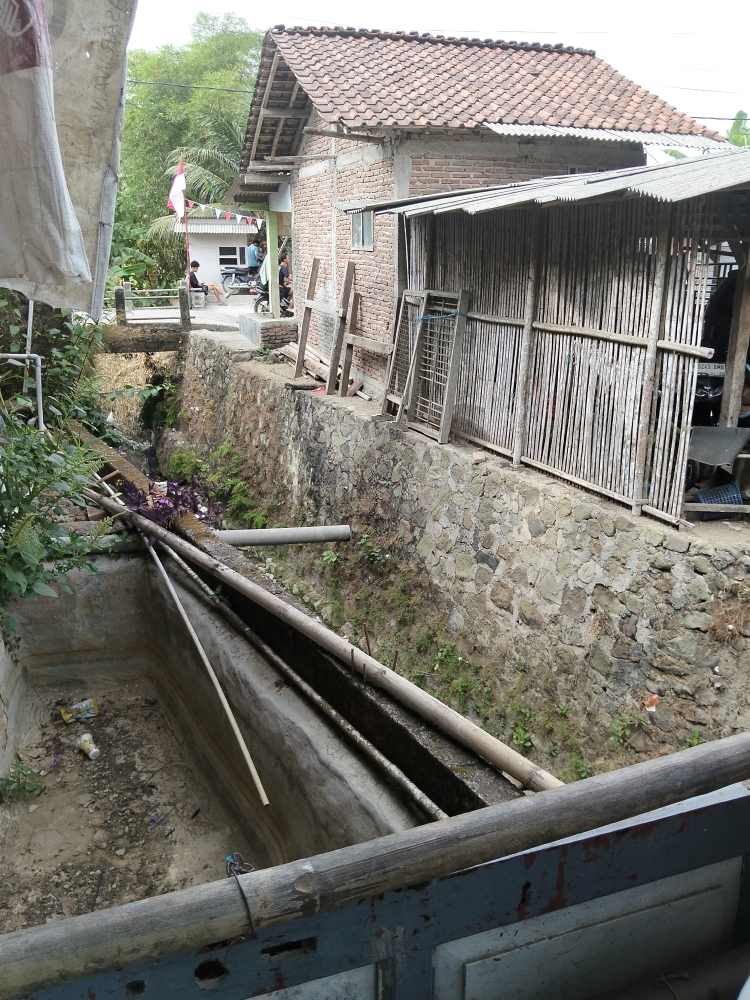 In Panimbang, Indonesia, a scene unfolds at street level, with a focus on a dry, concrete channel. The channel is littered with debris, including plastic waste and what appears to be a discarded cup, indicating a state of neglect. Along the top edge of the channel, several wooden beams and pipes are visible, suggesting some form of infrastructure or ongoing construction.
Beyond the channel, the environment transitions to a more built-up area. A stone wall forms a boundary, supporting a structure with a corrugated metal roof and walls constructed from vertical bamboo poles. Adjacent to this is a brick building with a red tiled roof. A few individuals are present in the background, near the bamboo structure and a small white building. One person sits on a railing, another stands near a motorcycle, and a couple more are seen in the distance. A red and white flag is displayed on a pole, and a string of small triangular flags is hung between buildings. The overall impression is of a residential or semi-commercial area characterized by simple construction and a somewhat unkempt appearance. The presence of vegetation, including trees and plants, suggests a tropical environment.