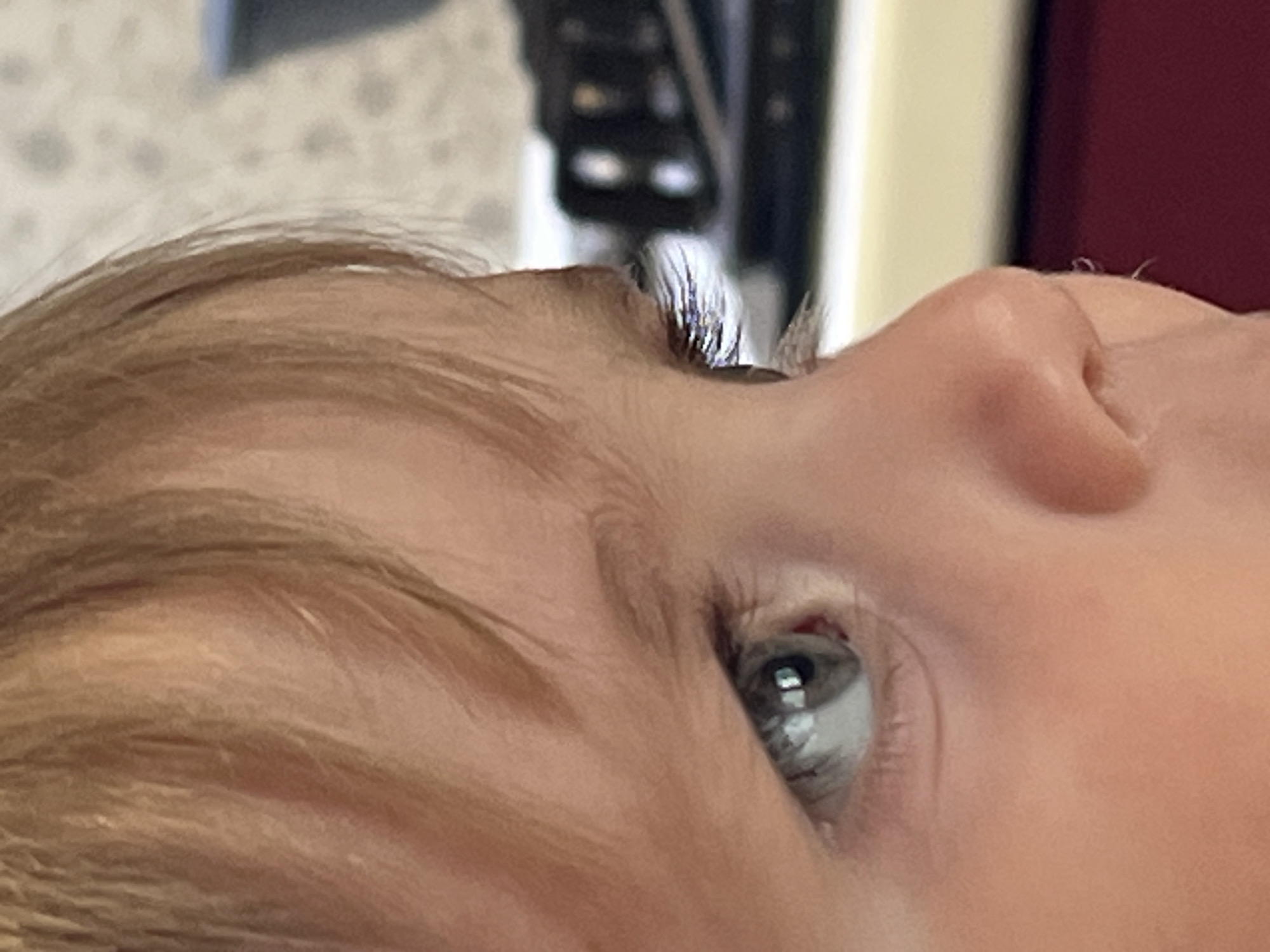 A close-up shot shows the face of a child with light brown hair and a blue eye. The child is looking upward. Only part of the nose and forehead are visible. The background is blurred and indistinct, but it appears to contain light-colored walls and dark objects. There is no information to suggest that this photo was taken in the city of Noviy, Russia.