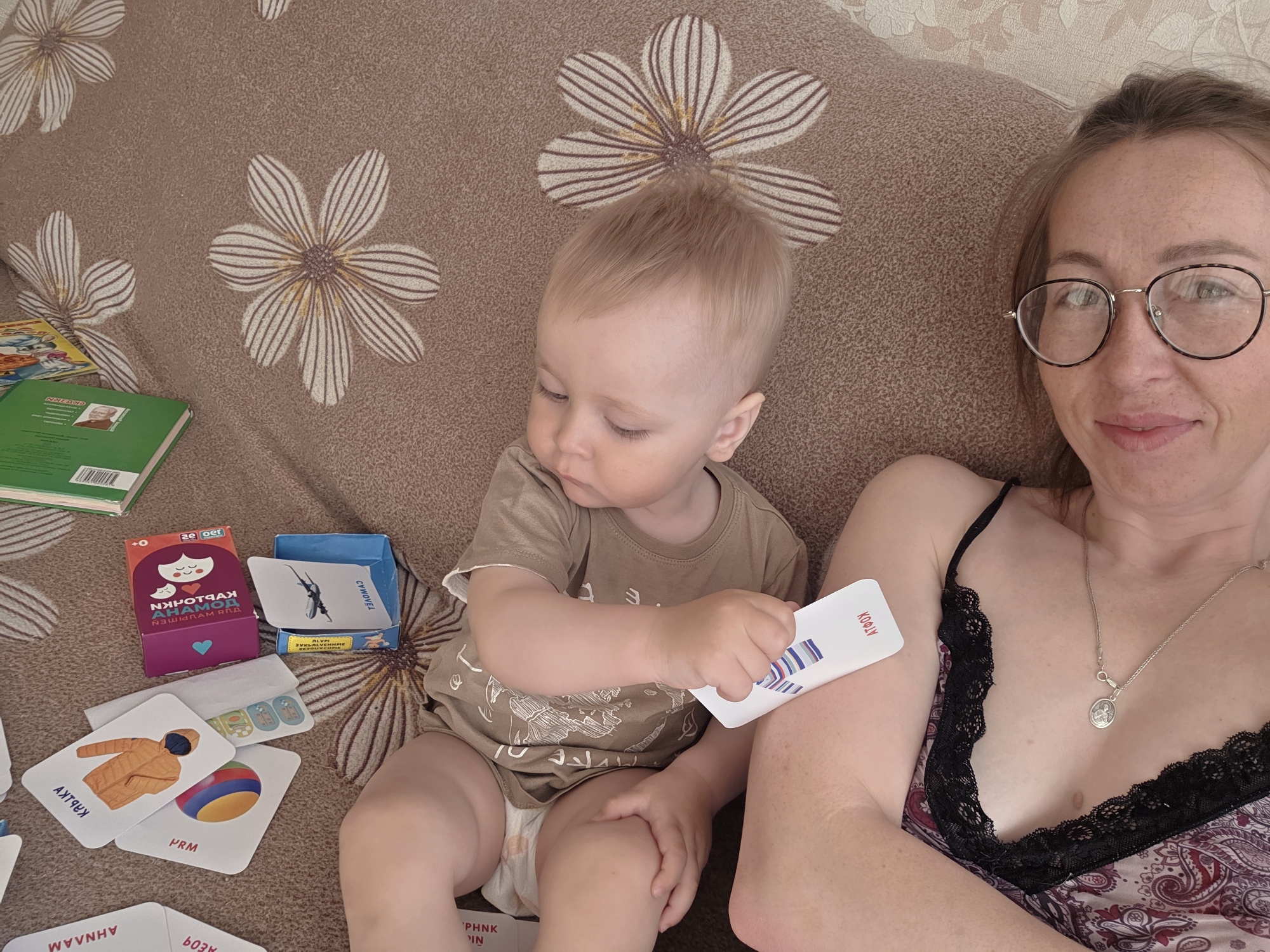 Here is a forensic description of the image:
A woman and a toddler are seated on a patterned sofa. The woman is fair-skinned and wearing glasses, a paisley-print top with black lace trim, and a silver necklace. She is looking at the camera. The toddler has blond hair and is wearing a brown t-shirt and a diaper. The child is holding up a card with "KOAI" written on it. On the sofa next to the child are several items, including books, a purple box with a cartoon drawing, a blue box, and several flashcards with images and words in a foreign language. The sofa has a brown and white flower pattern.