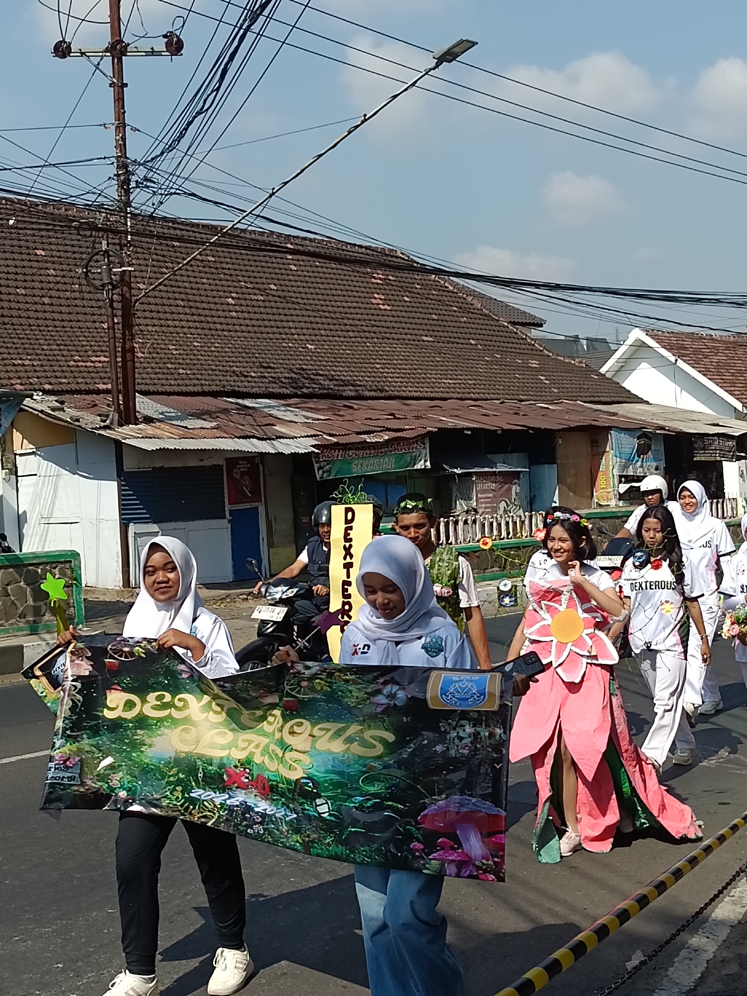 The image depicts a street scene in Kediri City, Indonesia, possibly during a parade or festival. Several young women, some wearing hijabs and others in uniforms, are walking on a paved road. One woman in the foreground holds a large banner with the text "Dexterous Class" displayed prominently, set against a backdrop of a vibrant, fantasy-like forest scene with mushrooms and stylized figures. Another woman is dressed in a costume resembling a large pink flower. Behind them, other participants and spectators are visible, including a person on a motorcycle. The surrounding environment includes buildings with tiled roofs, streetlights, and overhead electrical wires, indicative of an urban setting. The sky is clear and blue, suggesting a sunny day. The overall atmosphere appears festive and celebratory.