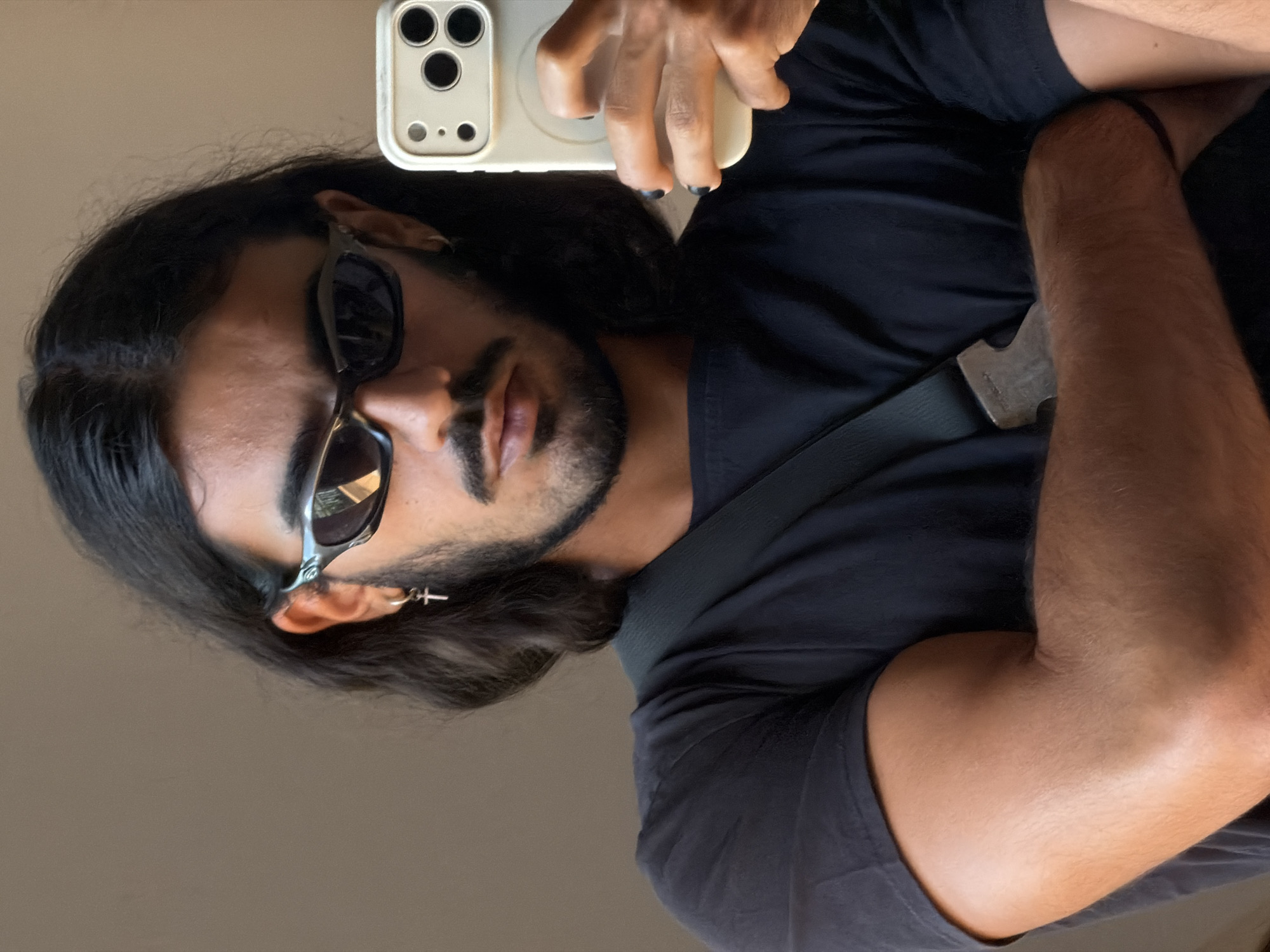 A mirror selfie captures a person with long dark hair, a neatly trimmed beard, and sunglasses. The individual, likely male, is wearing a plain dark-colored t-shirt and has a dark cross-shaped earring in their left ear. Their right hand, with dark-painted fingernails, holds a light-colored smartphone with a prominent triple-camera setup, seemingly an iPhone, which is used to take the photo. A dark strap, possibly from a bag, crosses their chest, and their left arm is flexed, revealing muscle definition. The background is a simple, light-colored indoor wall. The subject gazes towards the camera with a confident expression, partially obscured by the sunglasses. The scene depicts a casual self-portrait taken indoors, possibly in North Kuta, Indonesia. No visible text is present in the image.