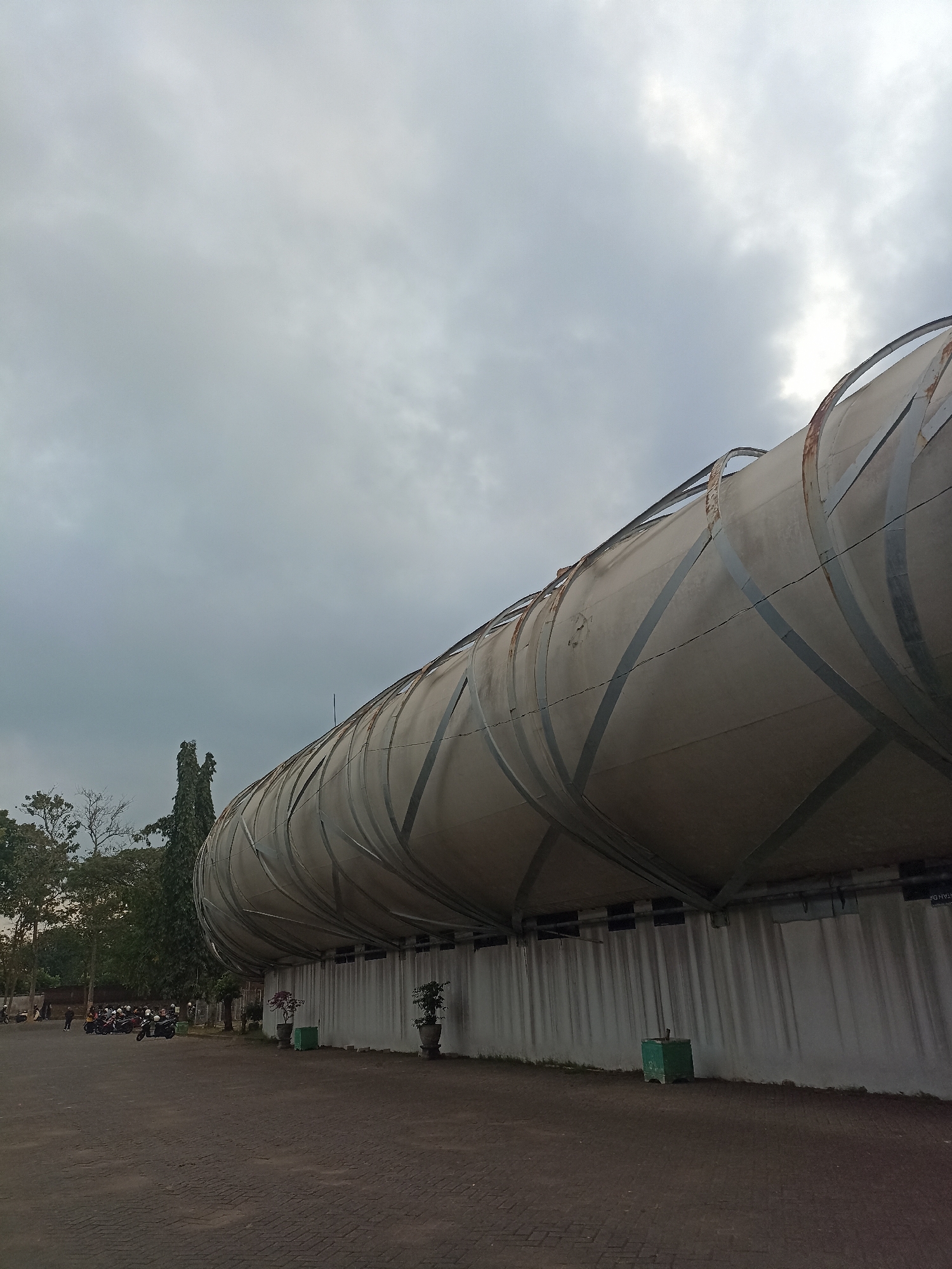 In Mojopanggung, Indonesia, a cylindrical structure with a distressed, metallic outer shell curves towards the right of the frame under a cloudy sky. The structure is supported by a white, vertically ribbed base. A paved area in front of the structure features several motorcycles parked in a line, with a few individuals standing near them. Trees and foliage are visible behind the structure to the left, with a tall, slender tree with dense foliage being a prominent feature. Two potted plants are placed in front of the base of the cylindrical structure. The overall scene depicts an outdoor environment with architectural elements and vehicles.
