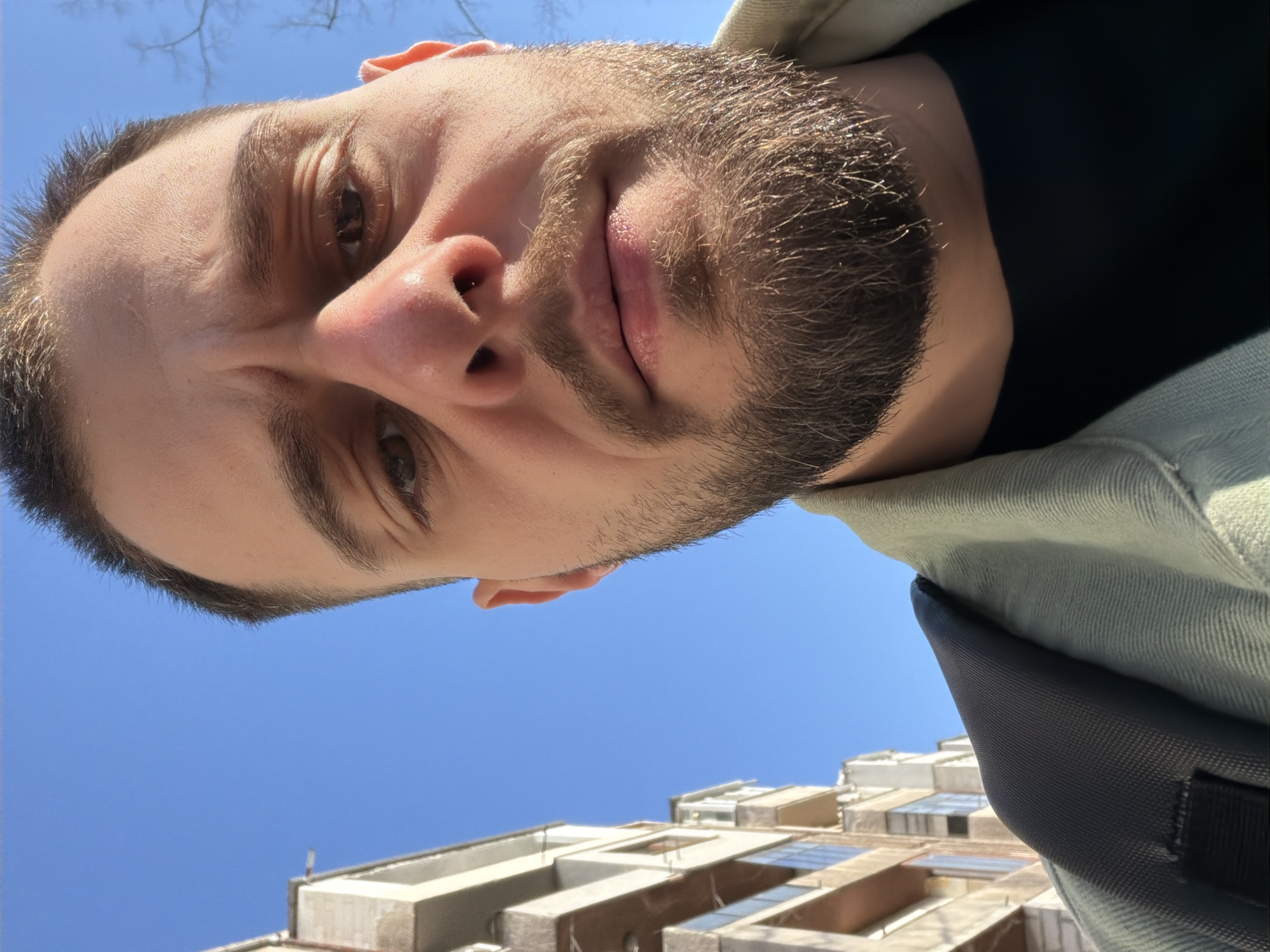The image shows a man looking directly down at the camera. He has short brown hair, light skin, and a beard. He is wearing a black shirt and a light green jacket, with a dark grey backpack strap visible on his right side. The background includes a clear blue sky and the top of a tall, multi-story building with balconies and multiple windows, appearing to be a residential apartment structure. The location is Plovdiv, Bulgaria.