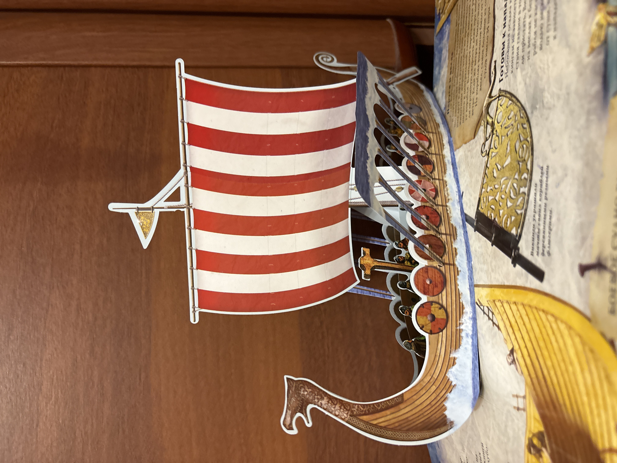 This is a close-up, slightly angled shot of a detailed pop-up book depicting Viking longships. The foreground features the front section of a Viking ship with a large red and white striped sail. Shields are visible along the side of the ship, and small figures of Vikings can be seen standing on deck. The ship is rendered with intricate details, including the wooden texture of the hull, the waves of the sea below, and a decorative dragon head at the prow.
Behind the primary ship, another pop-up element is visible, appearing to be another longship rendered in yellow. The background consists of a flat, dark brown surface, likely the cover or a page of the book itself, with some text in Russian. The text, partially visible and legible, includes phrases like "Готовы к напад" (Ready for attack) and mentions "Викинги" (Vikings), suggesting the book's theme. The lighting is bright, highlighting the vibrant colors and detailed artwork of the pop-up elements. The scene appears to be indoors, with the focus solely on the book's content.