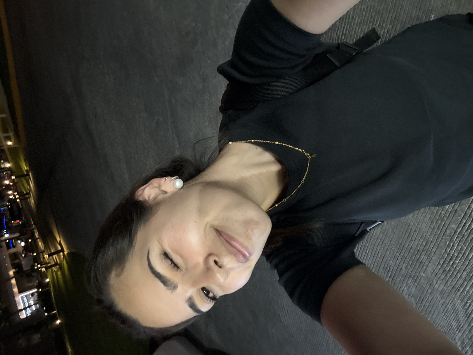 A woman lies on an outdoor surface in San Bernardino Tlaxcalancingo, Mexico. She wears a black shirt and has a gold necklace and pearl earrings. A building with lights is visible in the background. The ground appears to be paved.