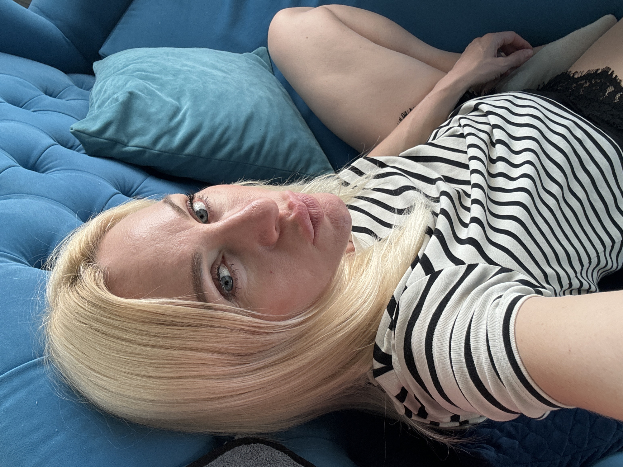 A blonde woman is lying on a blue couch, looking at the camera. She is wearing a black and white striped t-shirt and black lace shorts. Her hair is long and blonde, and she has blue eyes. She is pouting her lips. The couch is upholstered in a tufted blue fabric and has several matching blue pillows. The environment appears to be an indoor setting, likely a living room, with soft lighting. The image is a selfie, as evidenced by the angle and framing.