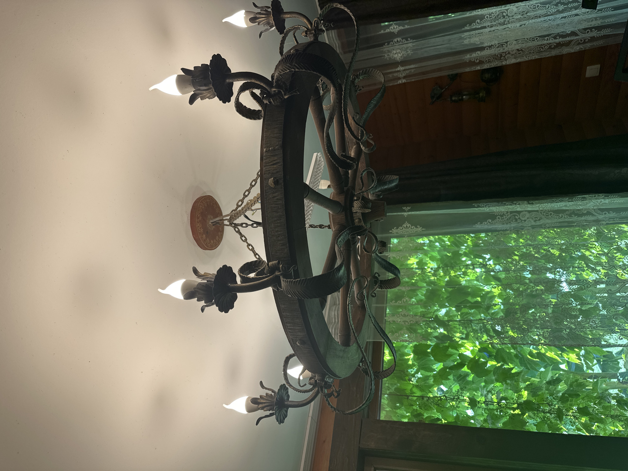 The image shows a rustic chandelier with six lights hanging from a ceiling. The chandelier is made of dark metal with twisted metal accents and appears to be a wheel-like design. Several of the light bulbs are illuminated, emitting a warm glow. The ceiling is white and appears to be textured.
To the right of the chandelier, a window is visible, covered by sheer white curtains with an ornate pattern. Through the window, lush green foliage is visible, suggesting the presence of trees or vines outside. The light filtering through the leaves creates a dappled effect. The background beyond the window appears to be a wooden interior, possibly a room in a house or a cabin.
The overall impression is one of a cozy and natural setting. The time of day is difficult to ascertain precisely, but the illuminated chandelier and the dappled light from the window could suggest late afternoon or early evening. The presence of the foliage and the rustic chandelier hint at a location with natural surroundings, possibly a dacha or a countryside home. No people are visible in the image, and no text is present.