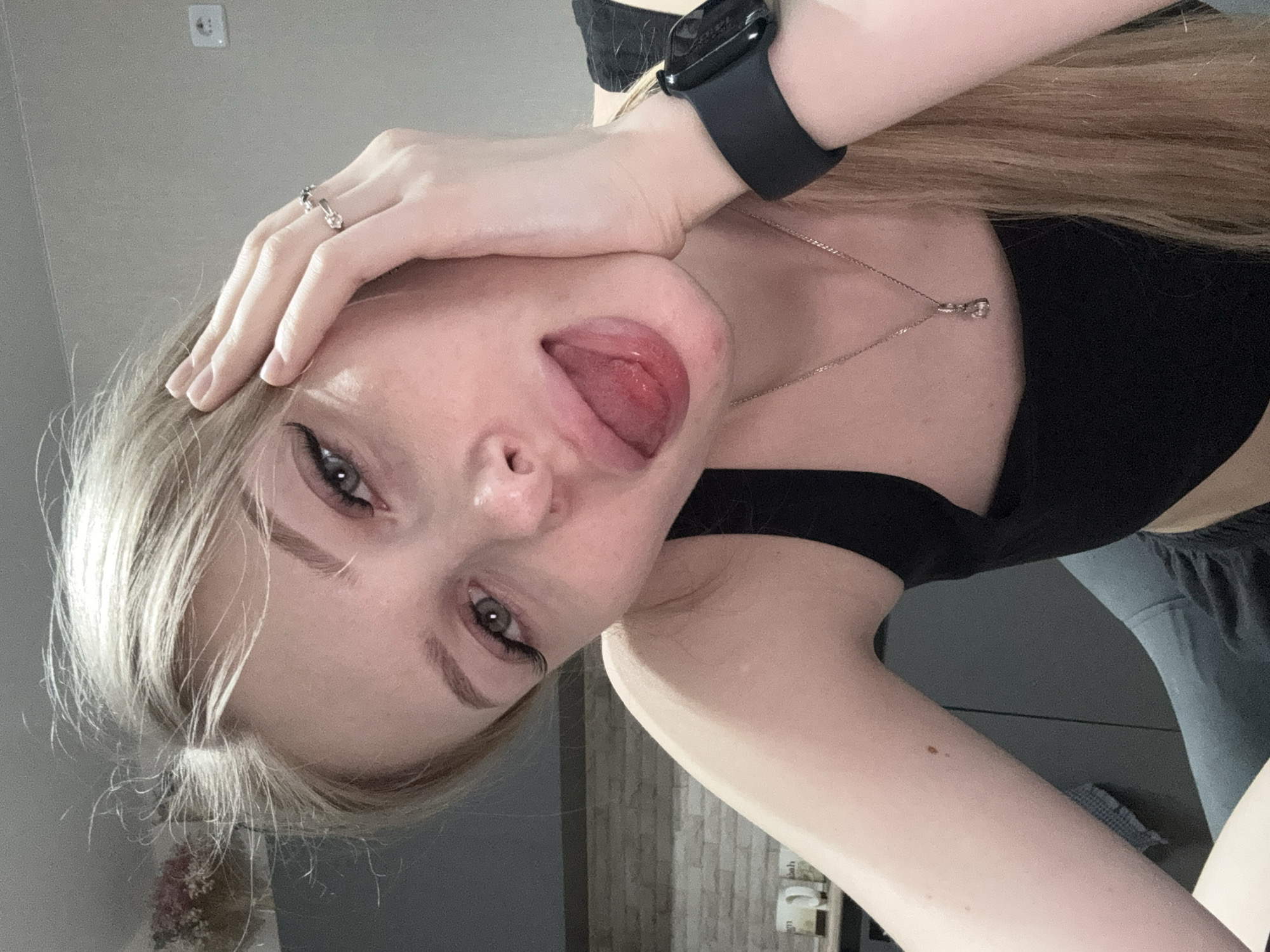 A light-skinned woman with blonde hair is seen at close range in a photograph oriented at an angle. The woman is wearing a black top, a silver necklace, and a black smartwatch. Her right hand is raised, fingers touching her forehead, and a ring is visible on one finger. She is sticking her tongue out.
The background includes a gray wall and a door with light brick detail. In the lower left corner, there is a partially visible object that appears to be a vase containing flowers. A switch is mounted on the wall in the upper left corner.
The location is likely indoors. The city of Perm, Russia is not discernible from this image.