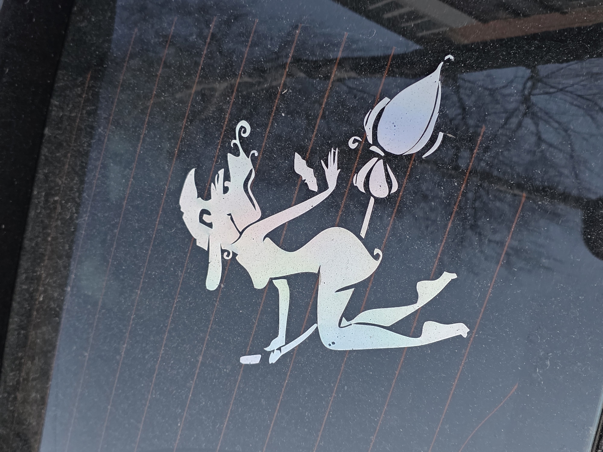 A white vinyl sticker of a cartoon witch riding a broom is adhered to the interior of a car's rear window. The sticker depicts a stylized, scantily-clad witch with pointy ears and wild hair, grinning while kneeling on a short broom. The broom's end resembles a flower or bulb. The window is tinted and dusty, and features heating element lines. The location is possibly Chaikovsky, Russia. The background shows vague reflections of trees and a building.