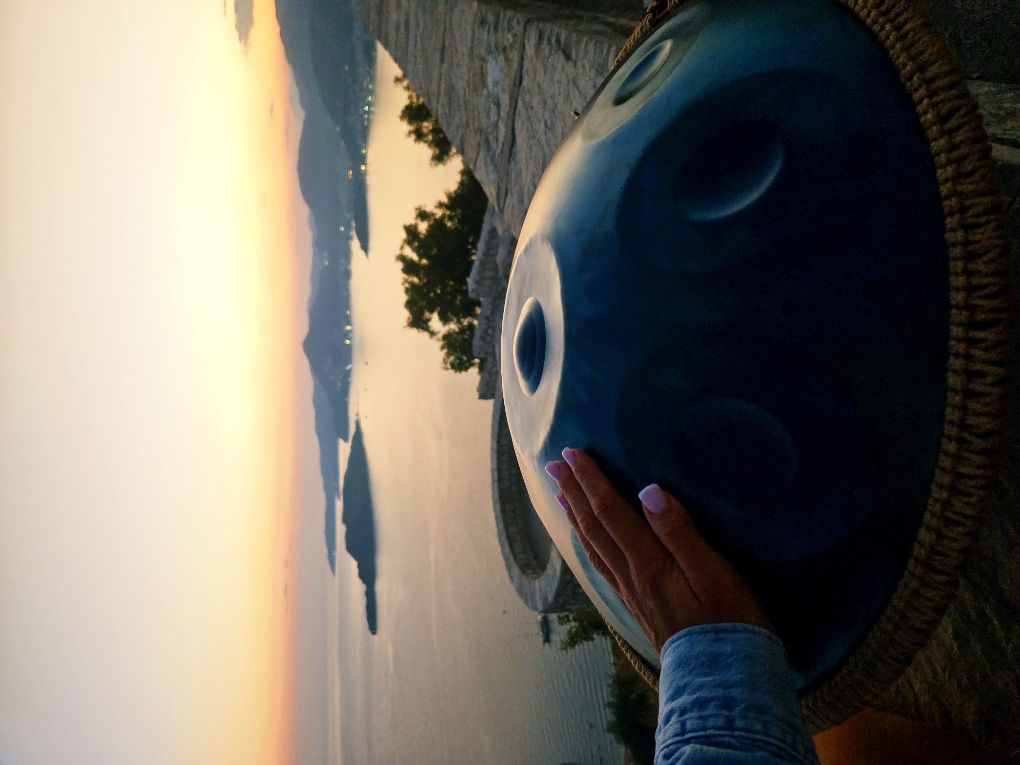 A hand with manicured fingernails rests on a dark blue handpan drum. The drum is positioned in the foreground, obscuring much of the background, which is a scenic view of the Adriatic Sea and the coastline of Sveti Stefan, Montenegro. The sky is a gradient of soft orange and yellow, indicating either sunrise or sunset. Islands and hills are visible in the distance, with lights suggesting a town or settlement. A stone wall and some foliage are also present in the scene. The overall mood is serene and peaceful, with the handpan suggesting a musical element to the natural beauty.