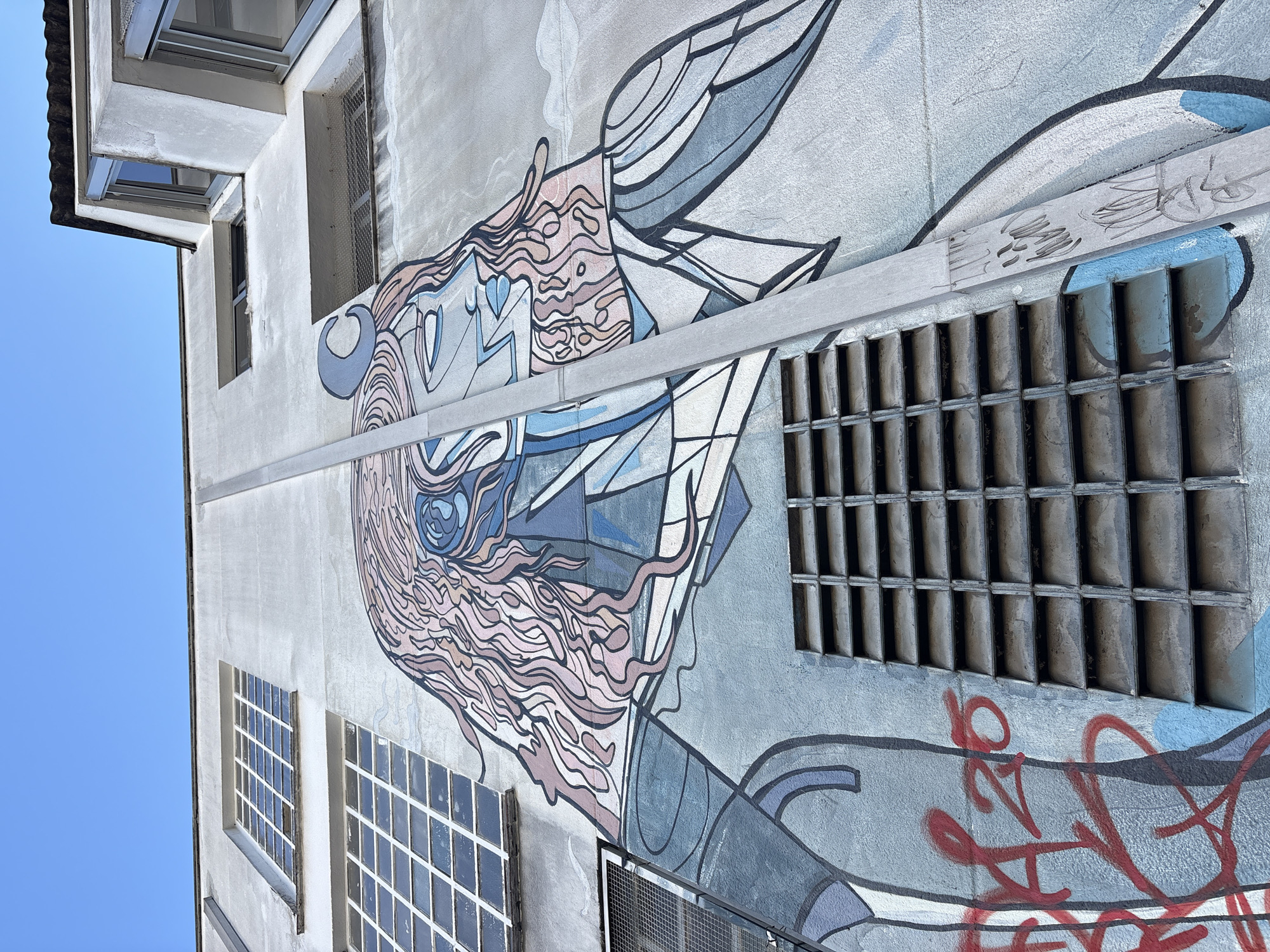 The image shows a mural on the side of a building in São Paulo, Brazil. The mural is a stylized depiction of a woman's face and upper body, with flowing pink hair and blue facial features. The woman has a crescent moon-shaped ornament on her head. The building appears to be made of concrete and has several windows. There is graffiti on the lower left side of the mural, in red. A vertical metal pipe runs down the center of the mural, obscuring part of the image. The sky above is clear and blue.