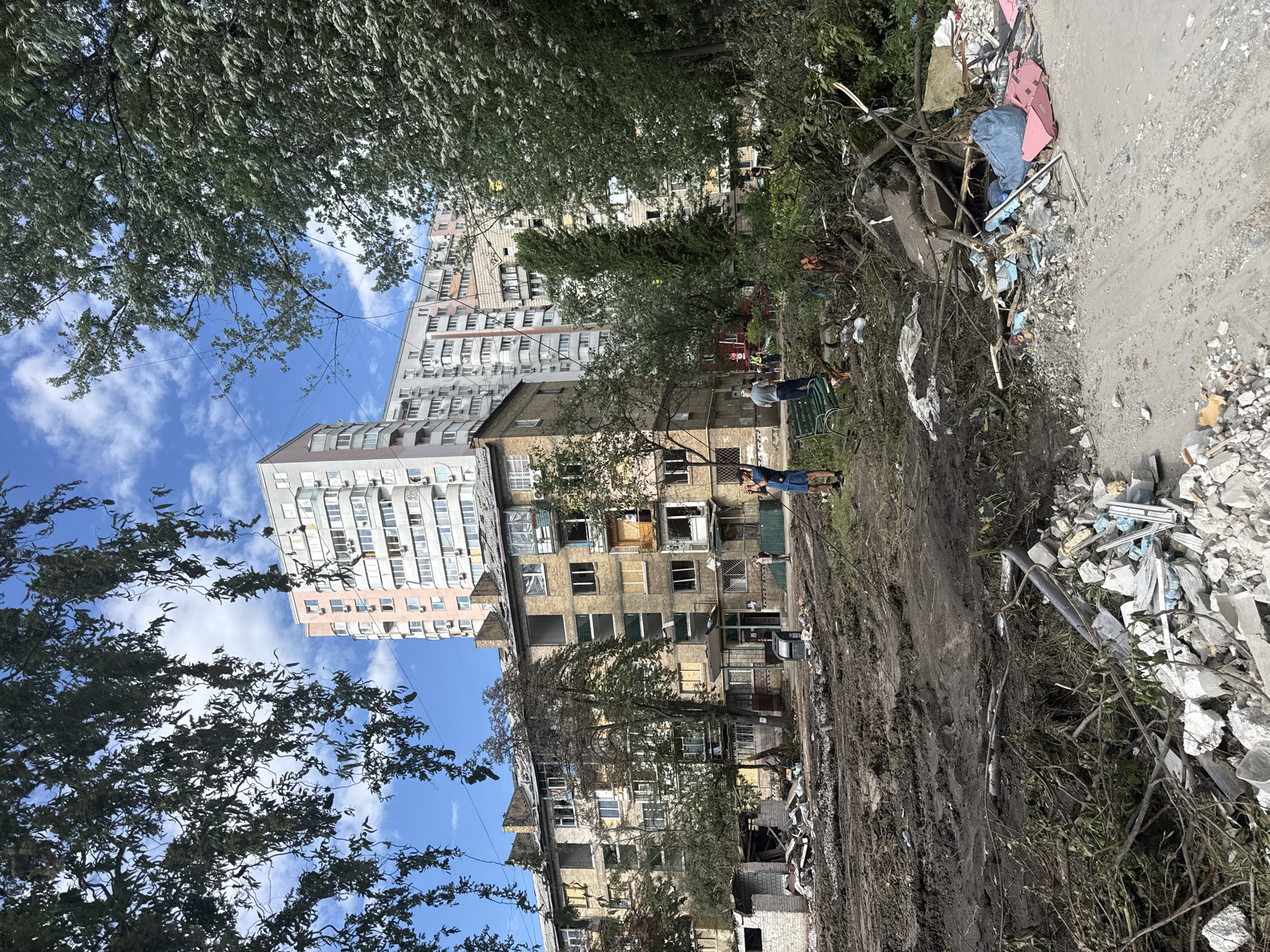 The image depicts the aftermath of destruction in Kyiv, Ukraine. In the foreground, a muddy, disturbed area shows tire tracks and debris, including broken concrete, branches, and plastic bags. A pile of construction waste and debris, including pink insulation foam and a blue plastic bag, sits at the edge of the muddy area.
Further back, a damaged apartment building with brick facade and broken windows stands under a blue sky with scattered clouds. Many windows are boarded up or have lost their glass. Adjacent to this building is a row of taller, modern apartment buildings with many windows and air conditioning units.
Several people are visible in the scene. In the mid-ground, two individuals stand near the damaged building, one appearing to be speaking with another. To the right, a person in a blue shirt and shorts holds a camera or similar device, possibly documenting the scene. Another person, wearing a dark t-shirt and shorts, is visible in the background.
The environment is urban, with residential buildings and trees. The overall impression is one of recent damage and ongoing cleanup or assessment efforts.