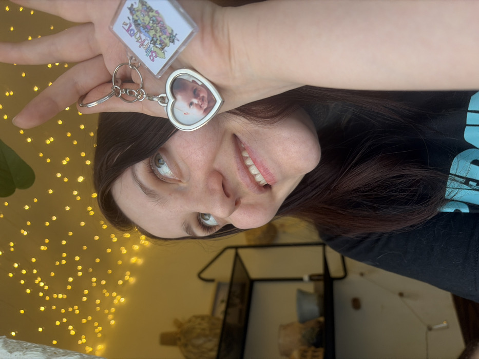 A woman smiles, holding two keychains up. One keychain is square, the other is heart-shaped with a baby's face inside. She has long brown hair and fair skin. Soft yellow fairy lights illuminate the background, which includes a shelf with decorative items. She wears a black t-shirt with blue writing.