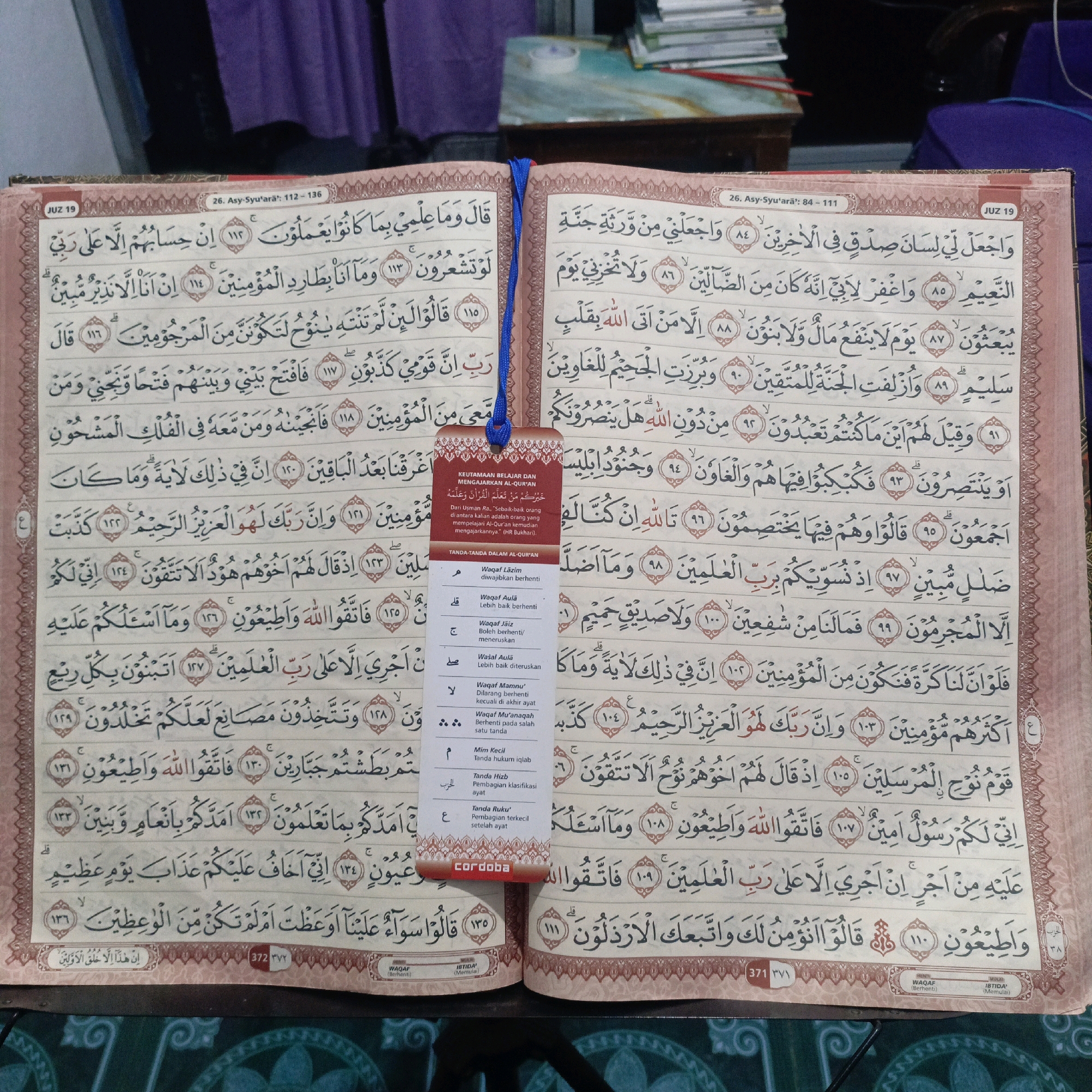 The image shows an open Quran on a table. The Quran is laid flat, with pages facing upwards. The left page displays verses 112-136 from Surah Asy-Syu'ara, and the right page displays verses 84-111 from the same Surah. The book is bound with a dark colored cover, and a blue bookmark is inserted between the pages. There are also small tags attached to the bookmark, one of which has "CORDOBA" printed on it, suggesting it is a religious book. The background is blurred, but appears to be an indoor setting with a soft purple fabric hanging behind the book. The lighting suggests it is daytime. The image is taken from a slightly elevated angle, looking down at the open book. There is no indication of the specific location within Salatiga, Indonesia.
